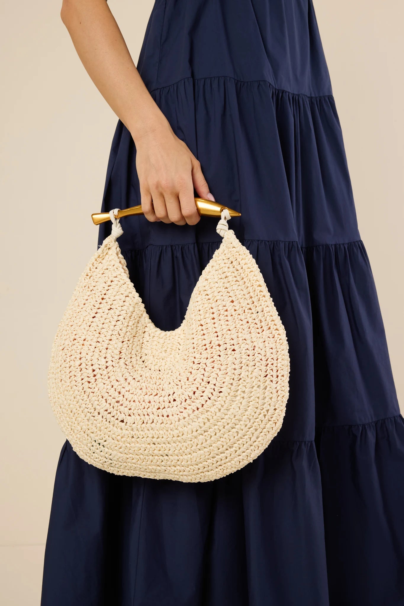 Person holding the Brielle Bag-Ivory, a crescent-shaped woven raffia handbag with a gold bar handle, ideal for stylish, versatile wear.