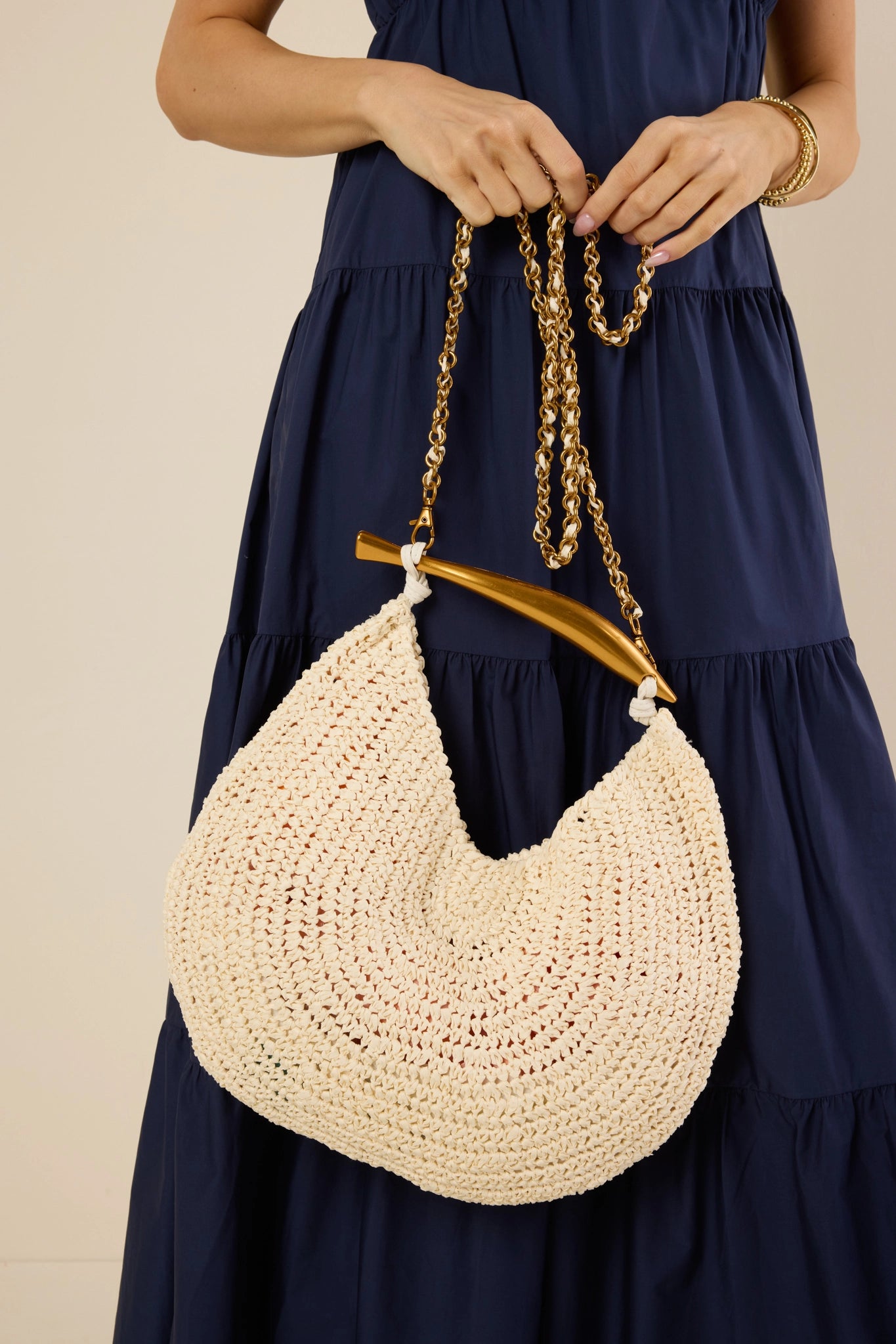 Person holding the Brielle Bag-Ivory, a crescent-shaped woven raffia bag with a gold-tone chain strap and sculptural accent, paired with a dark blue dress.