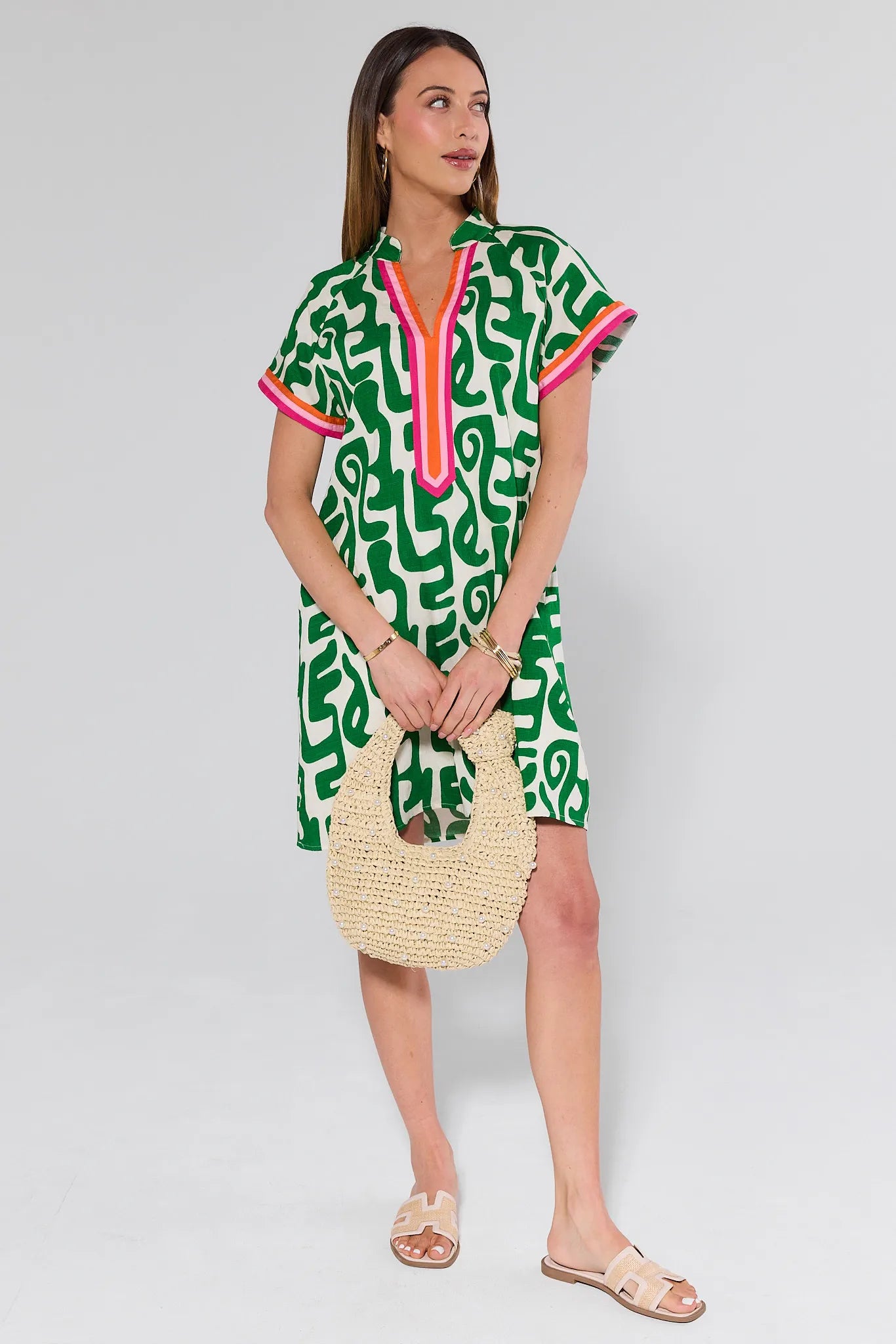 Woman wearing a green patterned dress with pink accents, holding a straw bag on a white background
