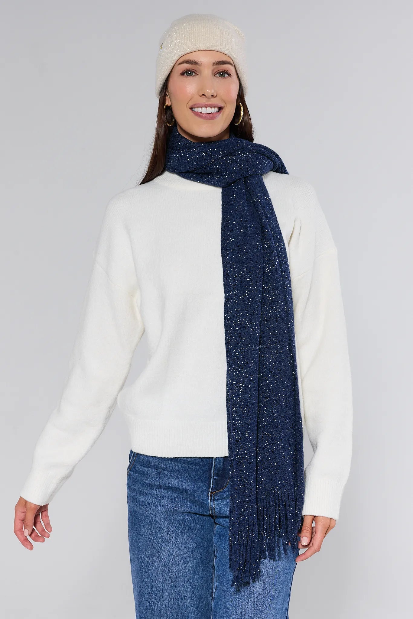 Woman wearing a white sweater, blue jeans, and a Karris Navy Fringe Scarf with a beige beanie on a gray background