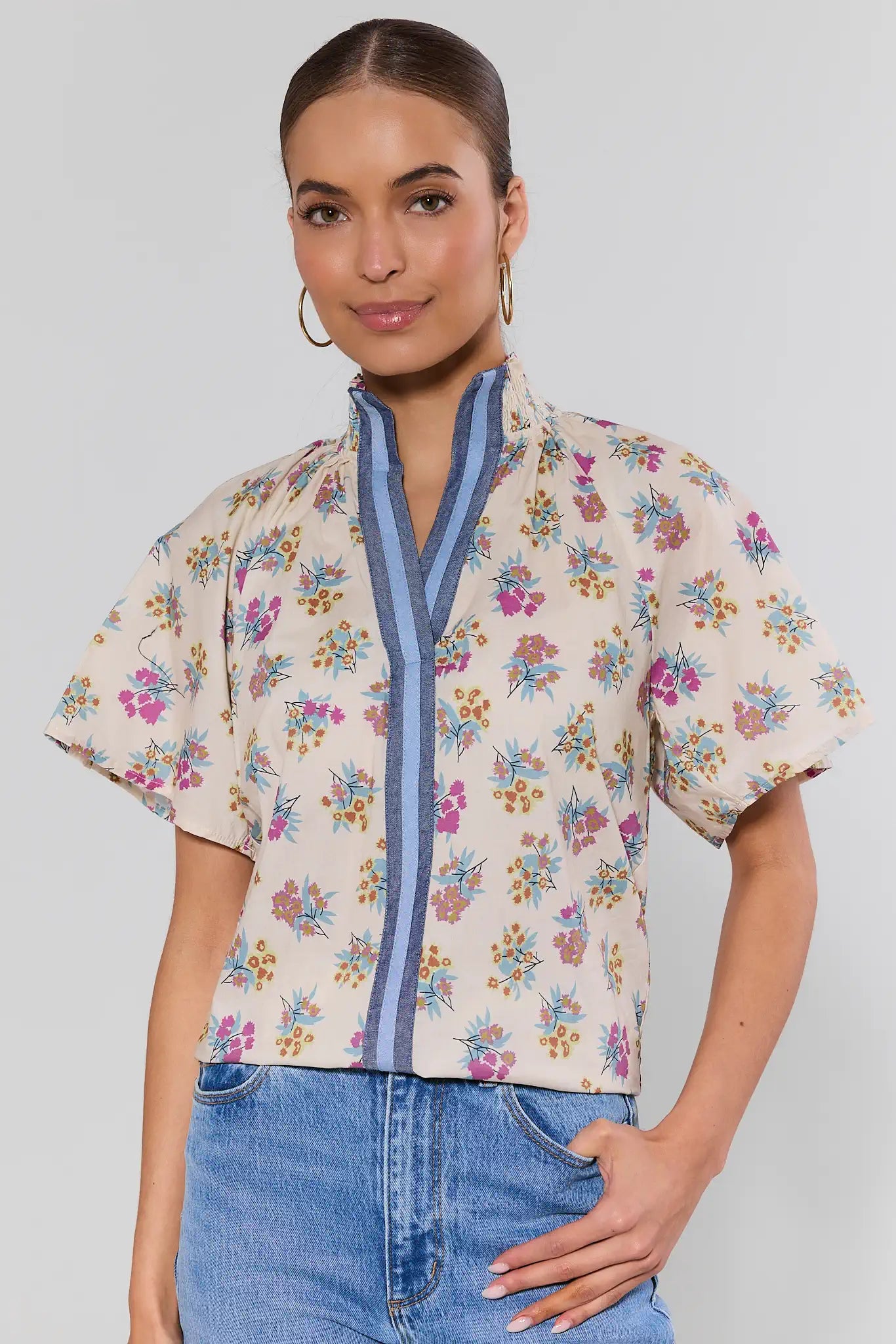 Model wearing Macey Cream Floral Print Top by THML, featuring short sleeves, a stand collar, and blue-striped trim, paired with high-waisted denim jeans.