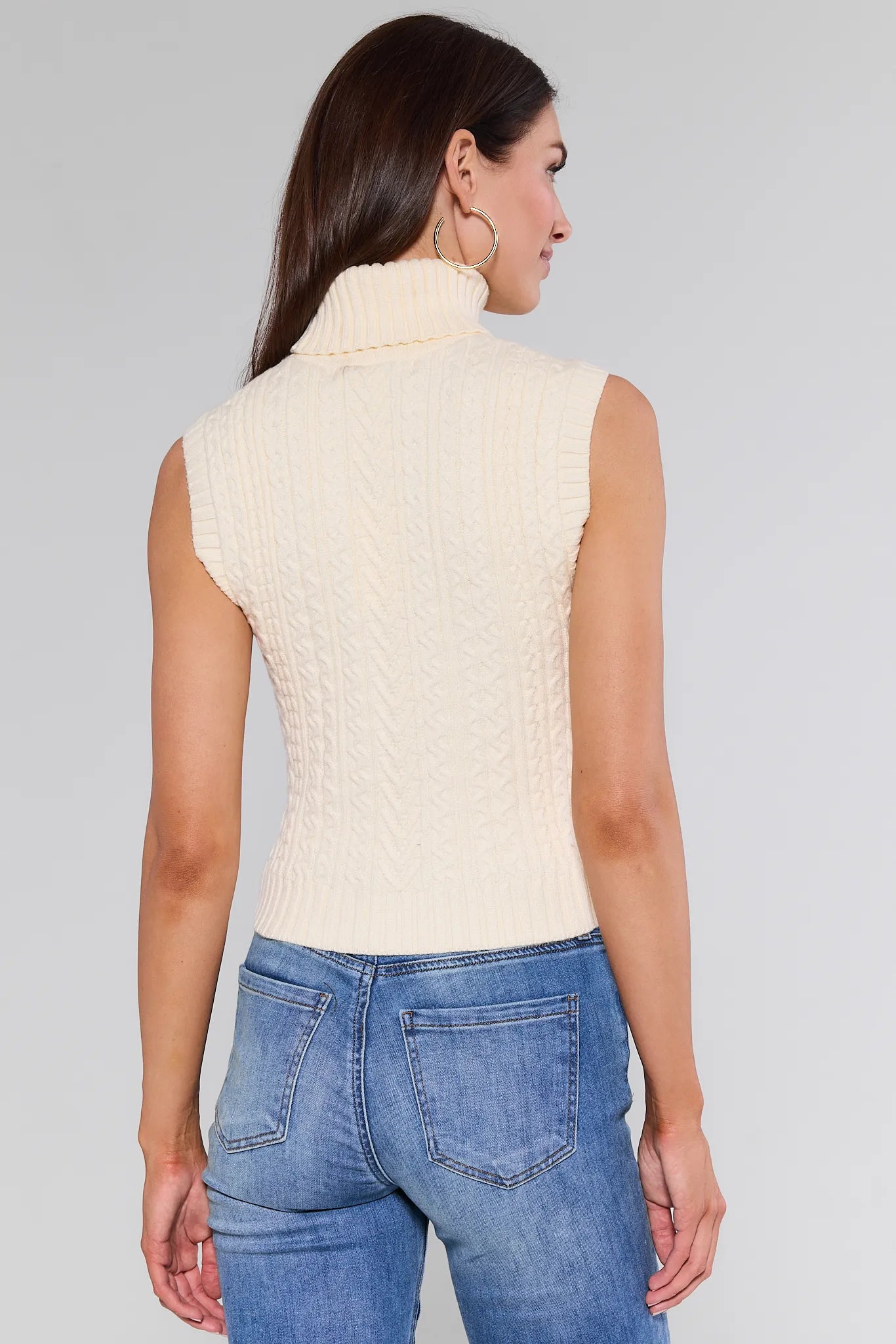 Woman wearing a cream sleeveless knitted Sienna Sweater and blue jeans against a gray background