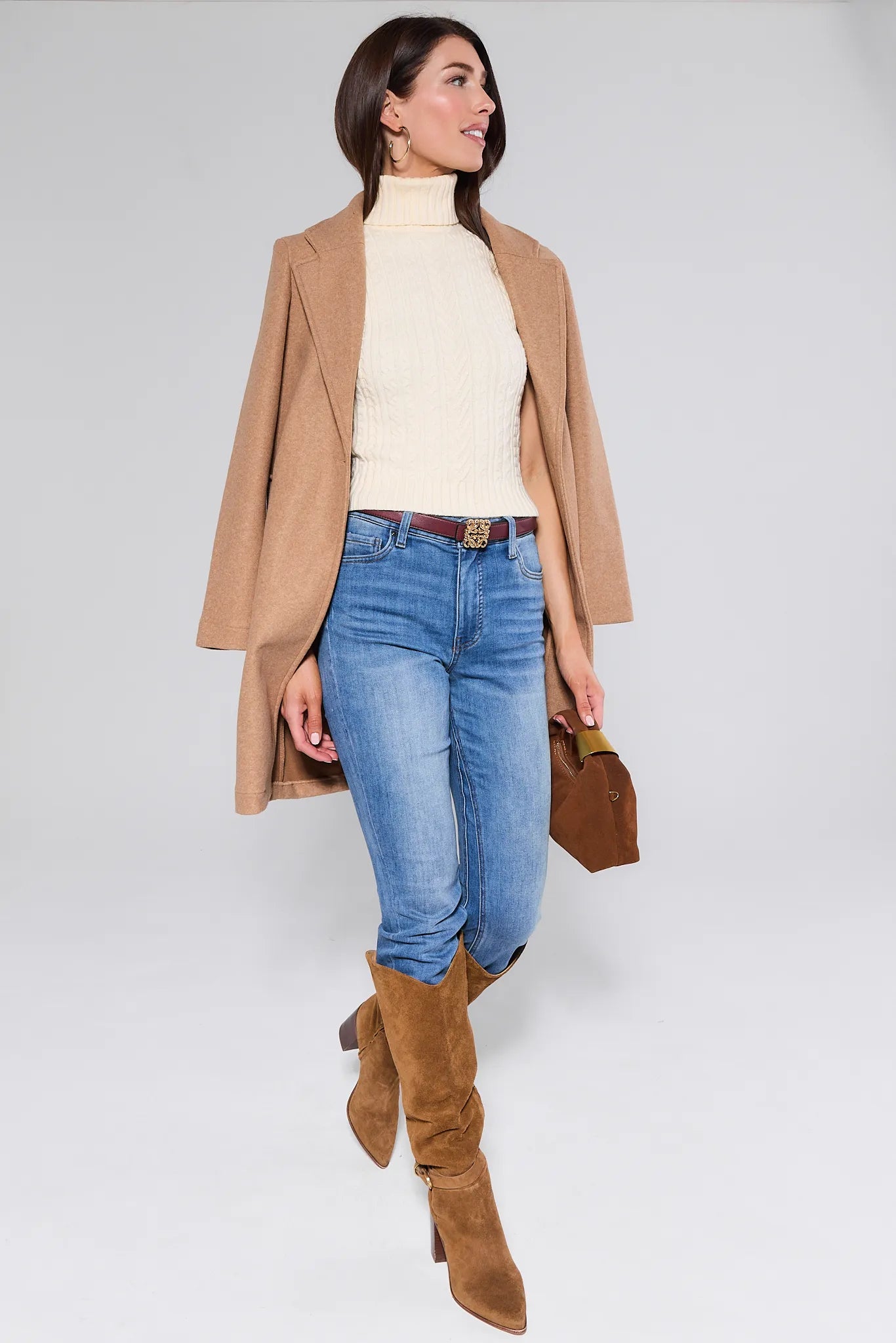 Woman wearing a tan coat, cream Sienna Sweater, blue jeans, and brown boots on a white background