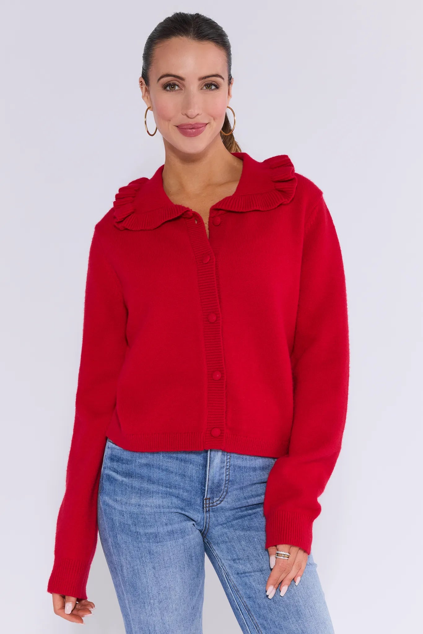 Woman wearing a red jordyn cardigan with a ruffled collar and blue jeans on a white background