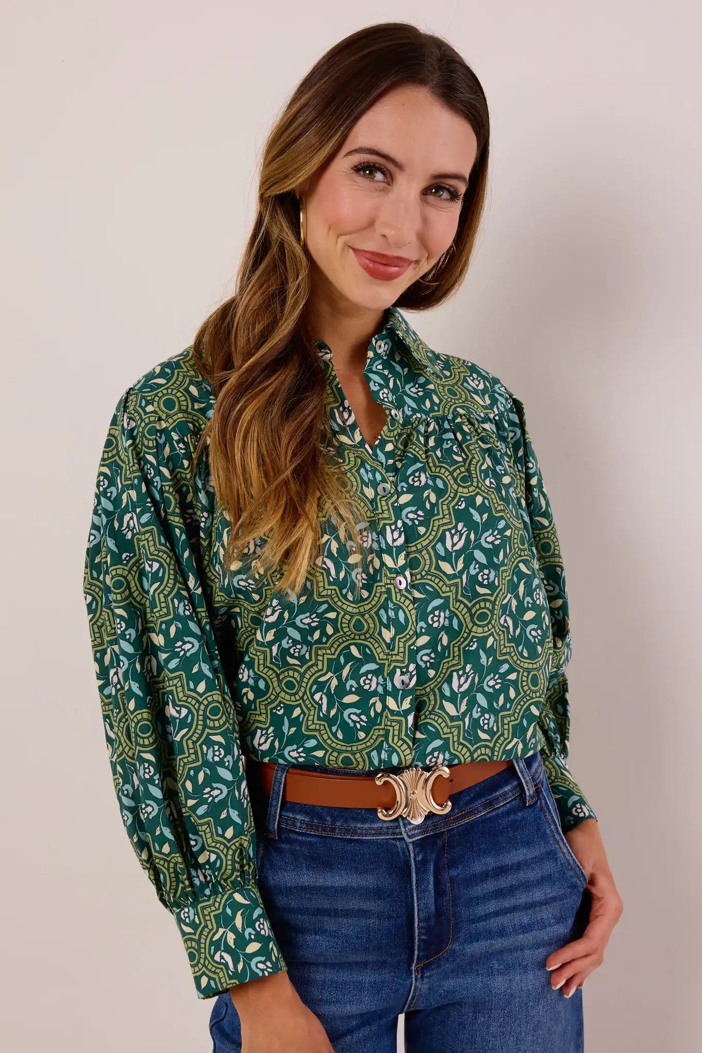 Young woman modeling the Trudy Olive Floral Top, featuring a geometric-floral print, button-front closure, and relaxed fit, paired with blue jeans and a brown belt.