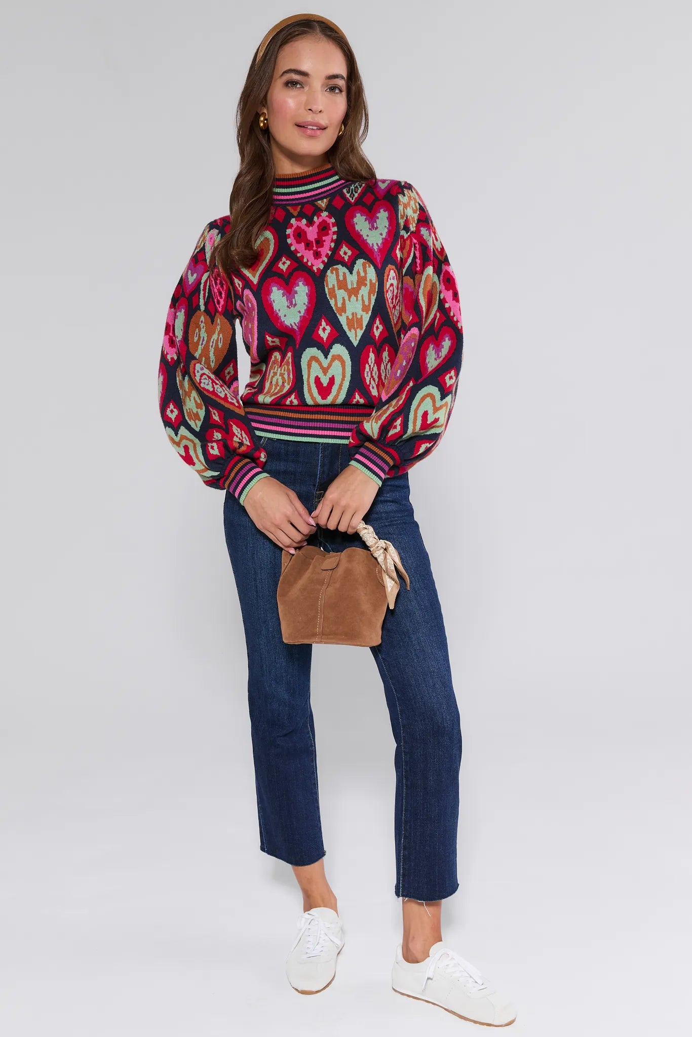 Woman wearing a colorful patterned Maddie sweater with hearts, blue jeans, and white sneakers on a white background.