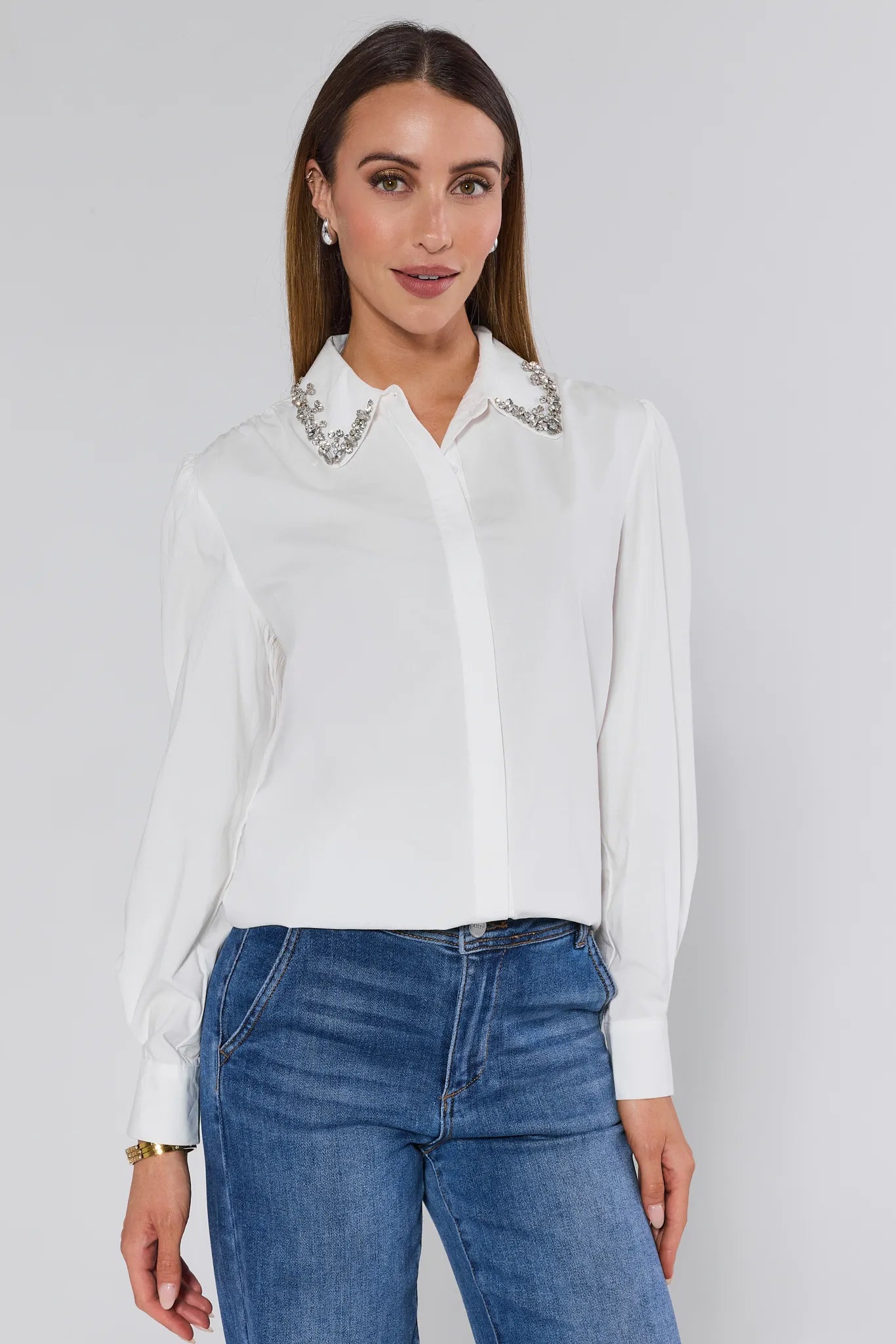 Woman wearing a white Celeste Top with embellishments and blue jeans on a plain background