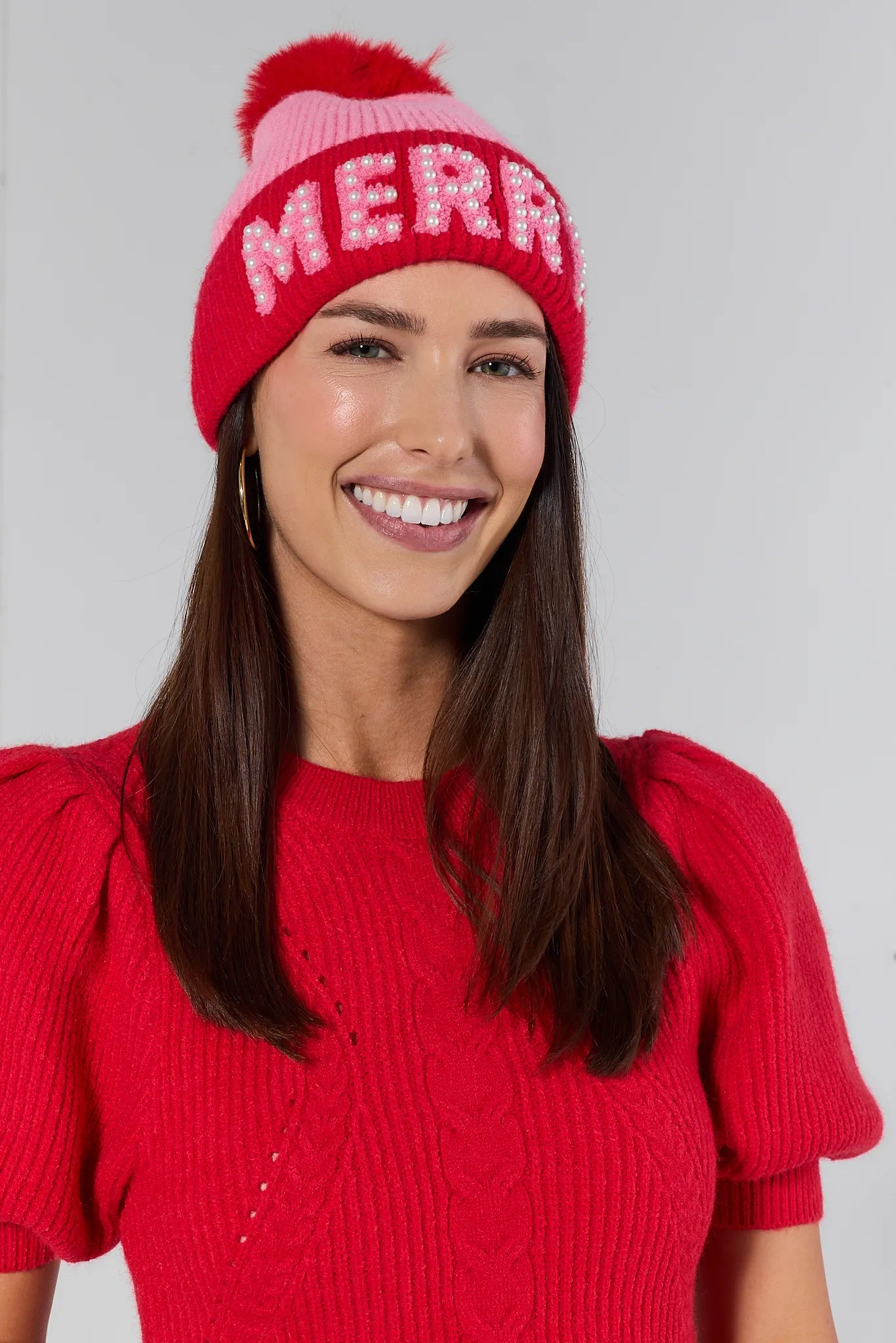 Woman wearing a red knit sweater and pink beanie with 'MERRY' on a plain background