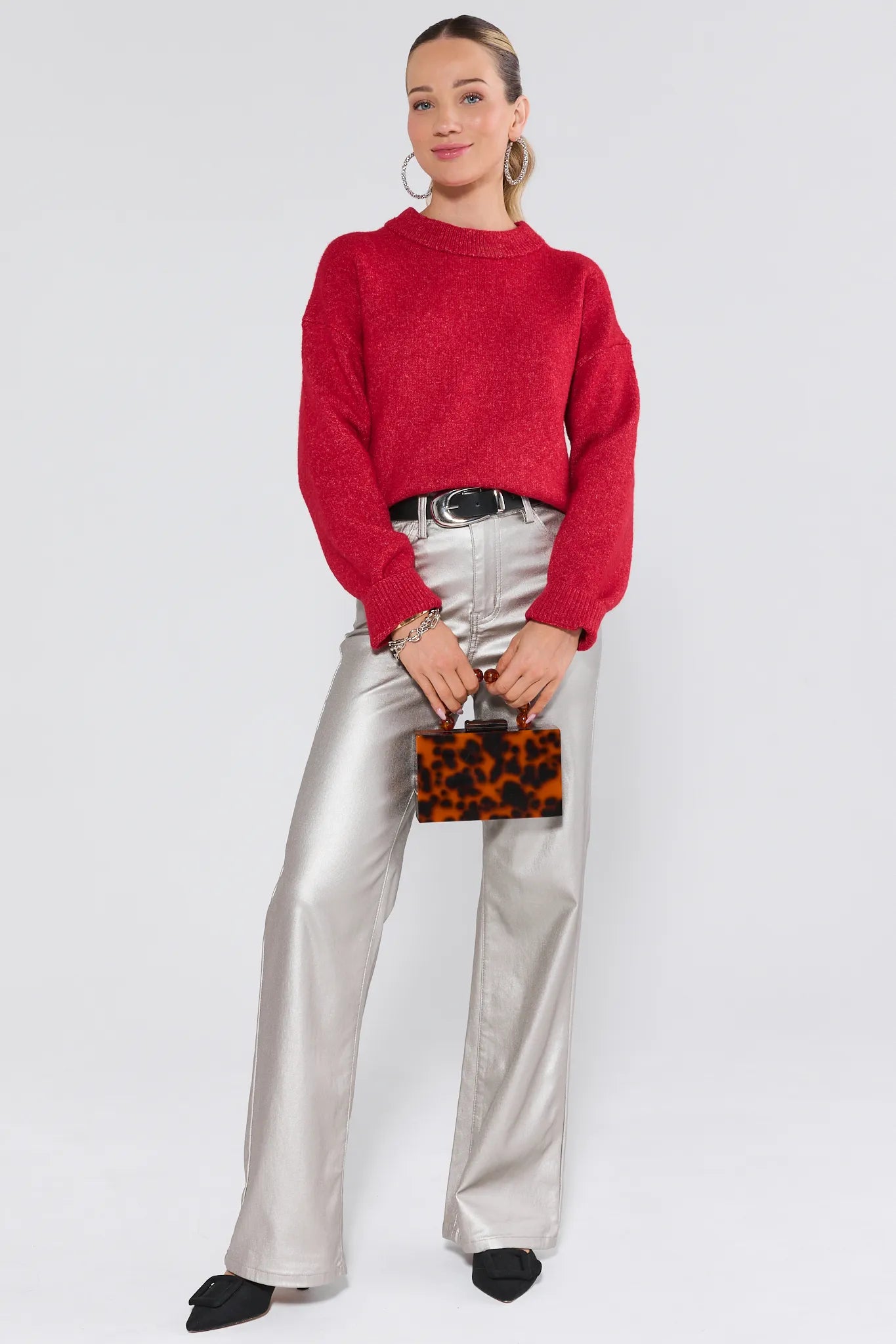 Woman wearing a red sweater and silver pants holding a leopard print clutch against a white background