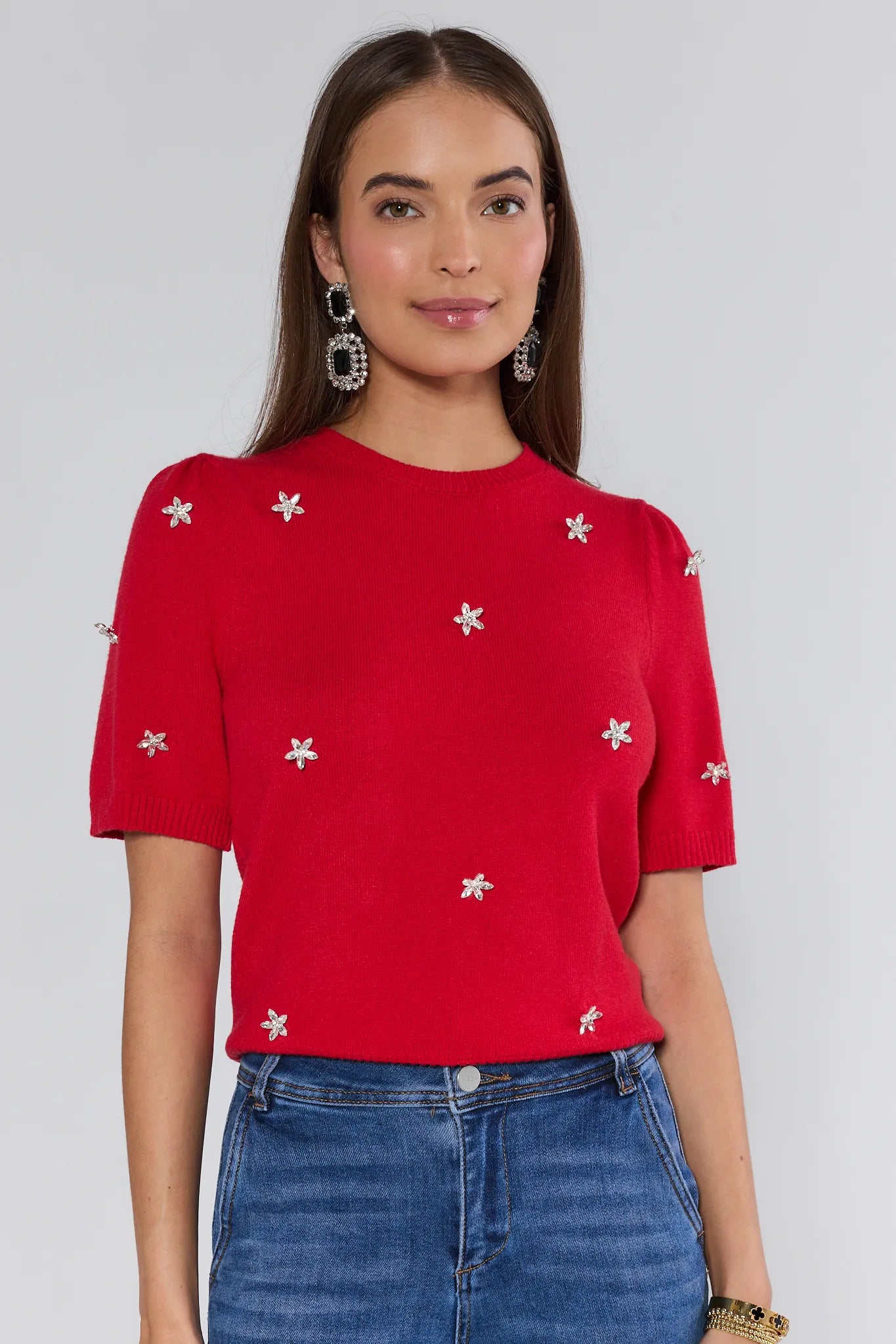 Woman wearing a red top with white floral patterns and blue jeans on a plain background