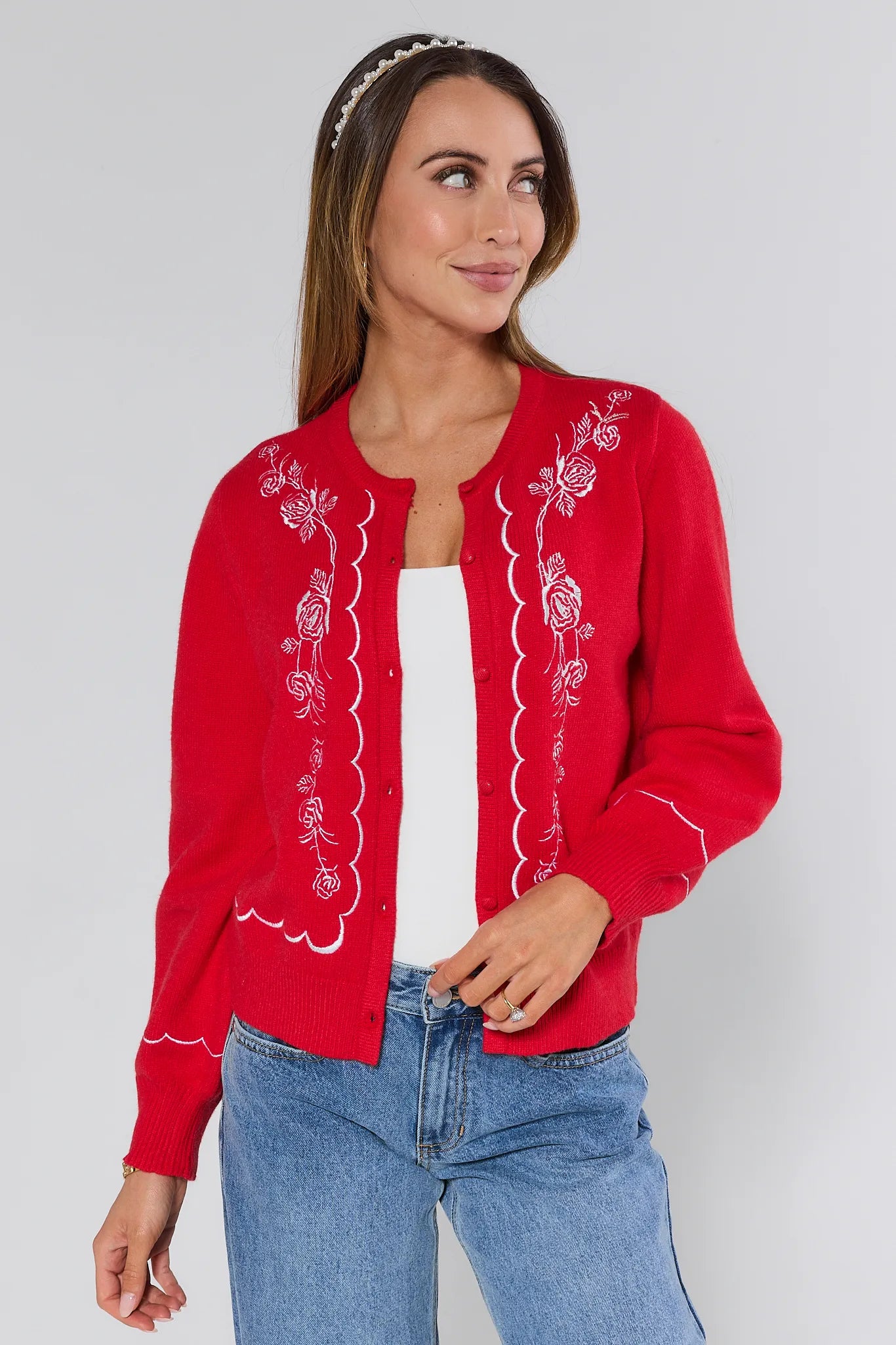 Woman wearing a red embroidered Ginger cardigan over a white top and blue jeans on a plain background