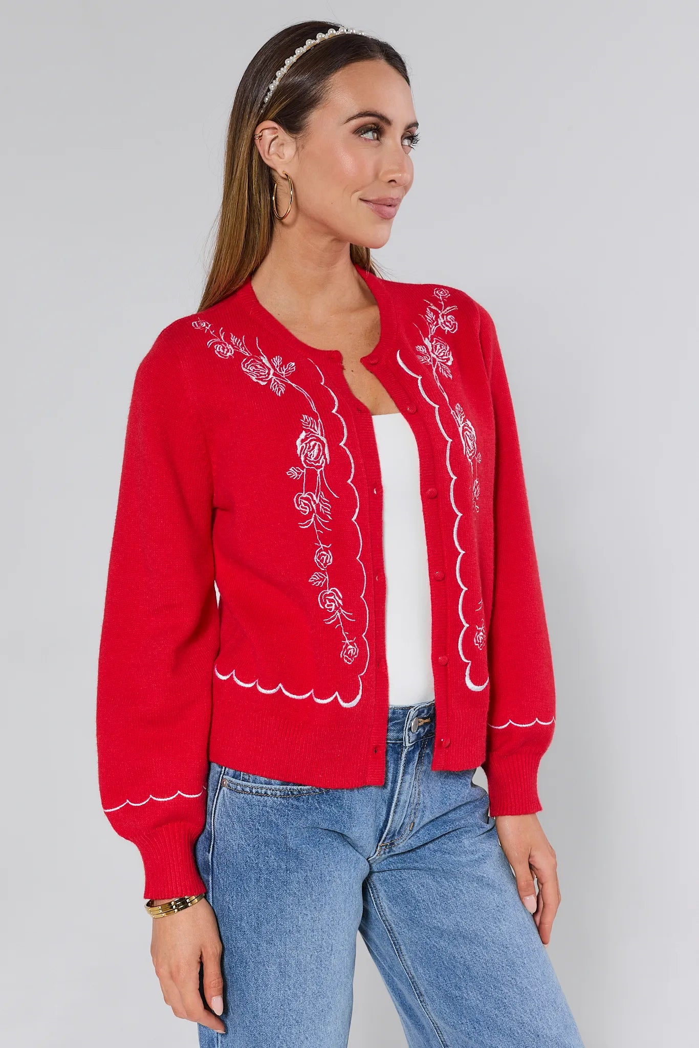 Woman wearing a red embroidered Ginger cardigan over a white top and blue jeans on a plain background