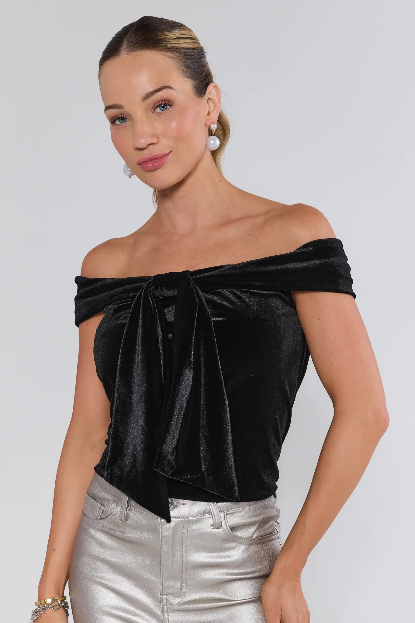 Woman wearing a black off-shoulder velvet top and silver pants on a white background