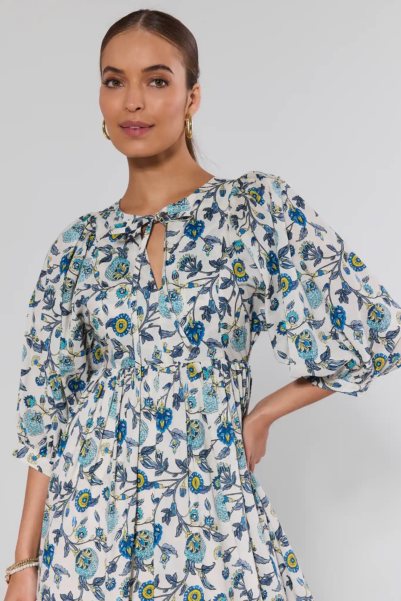 Woman wearing Paige Blue Floral Print Short Dress with puff sleeves, A-line silhouette, keyhole neckline, and gold hoop earrings, hand on hip, hair pulled back.