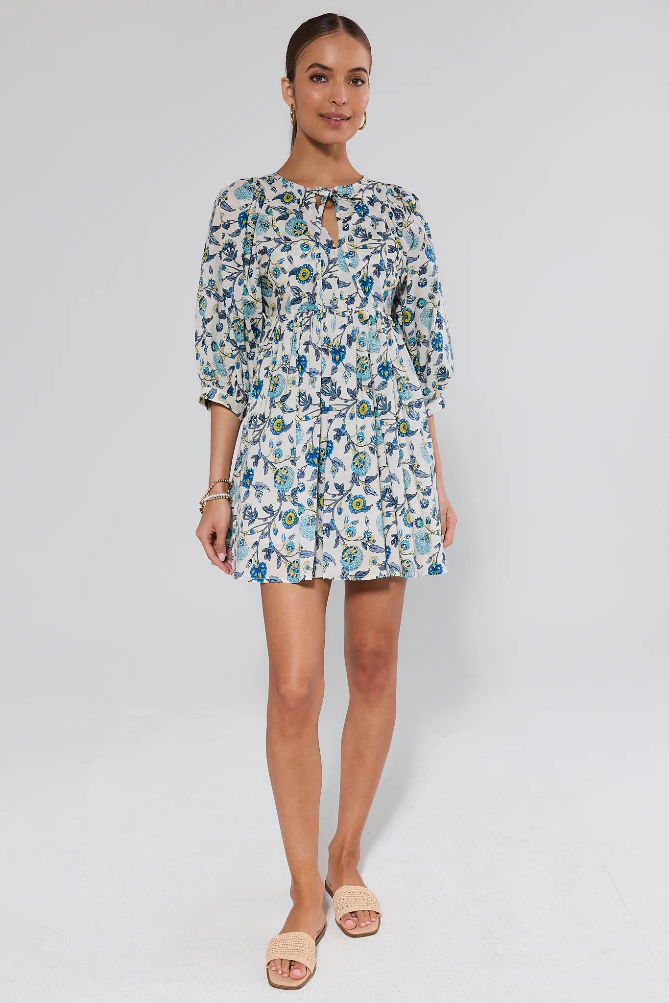Paige Blue Floral Print Short Dress worn by a dark-haired woman, featuring a keyhole neckline, tiered design, and puff sleeves, paired with beige woven sandals.
