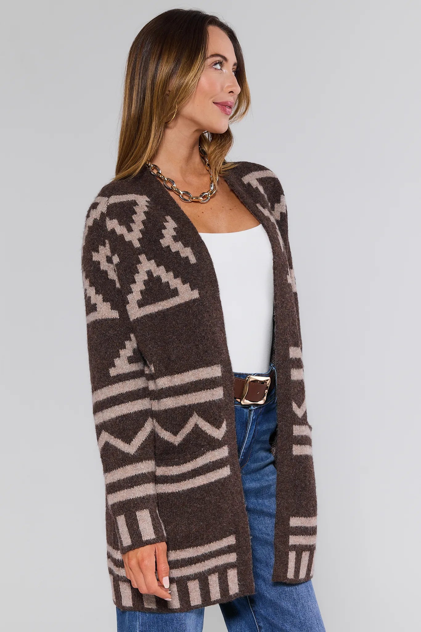 Woman wearing a brown patterned Helena Cardigan Brown over a white top and blue jeans on a gray background