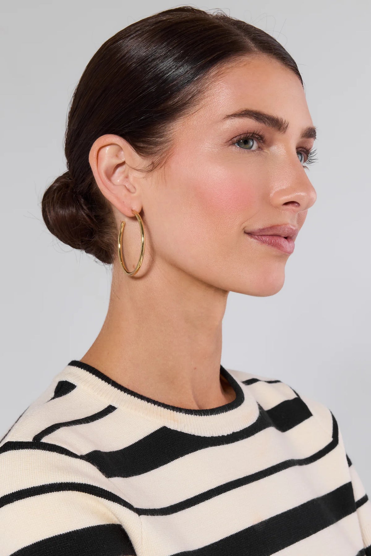 Woman wearing Leigh Gold Hoop Earrings, 2 in diameter, with a sleek, 14k gold-dipped finish, complementing her black-and-white striped top and natural makeup.