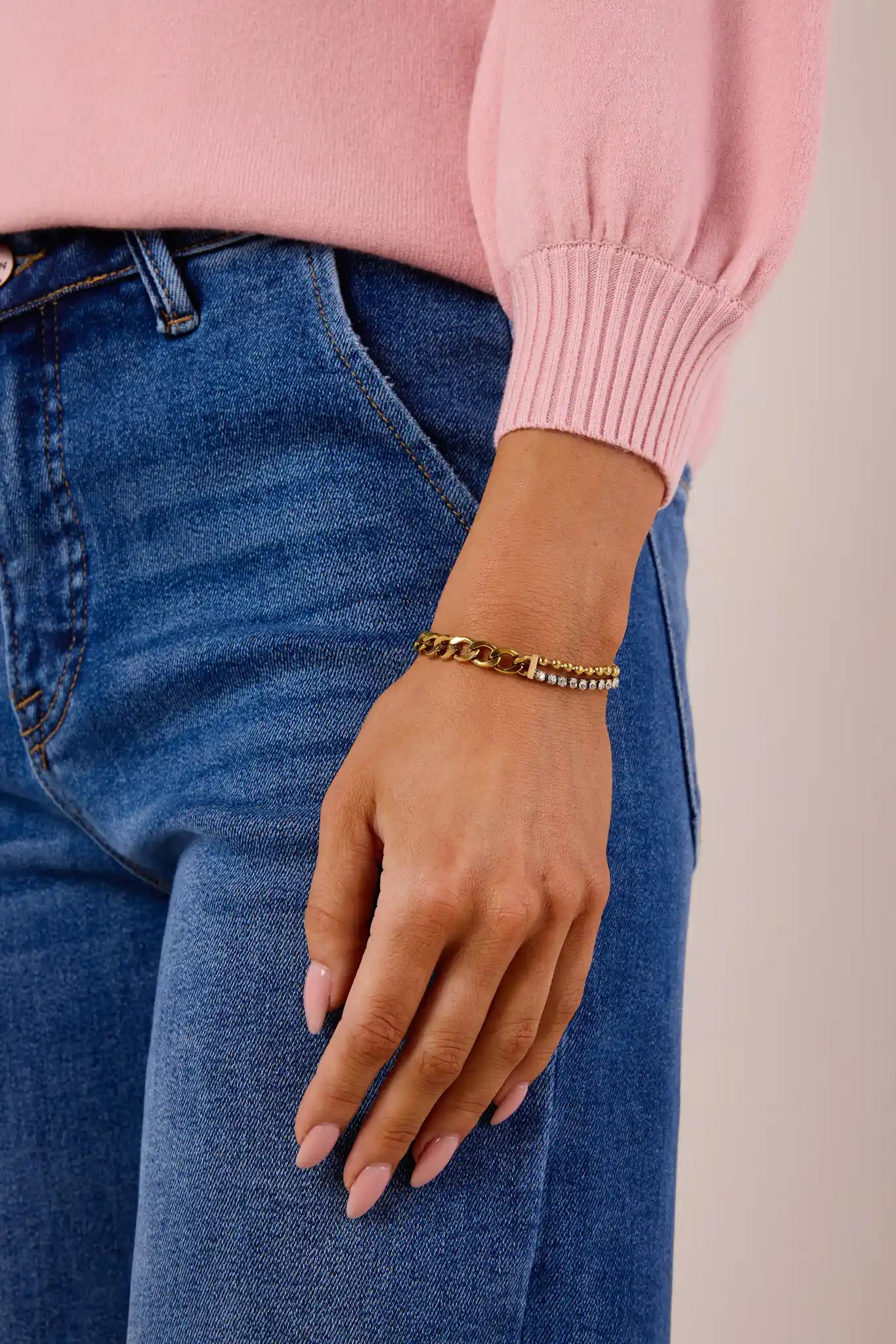 Person wearing a pink sweater and blue jeans with a gold bracelet on a neutral background