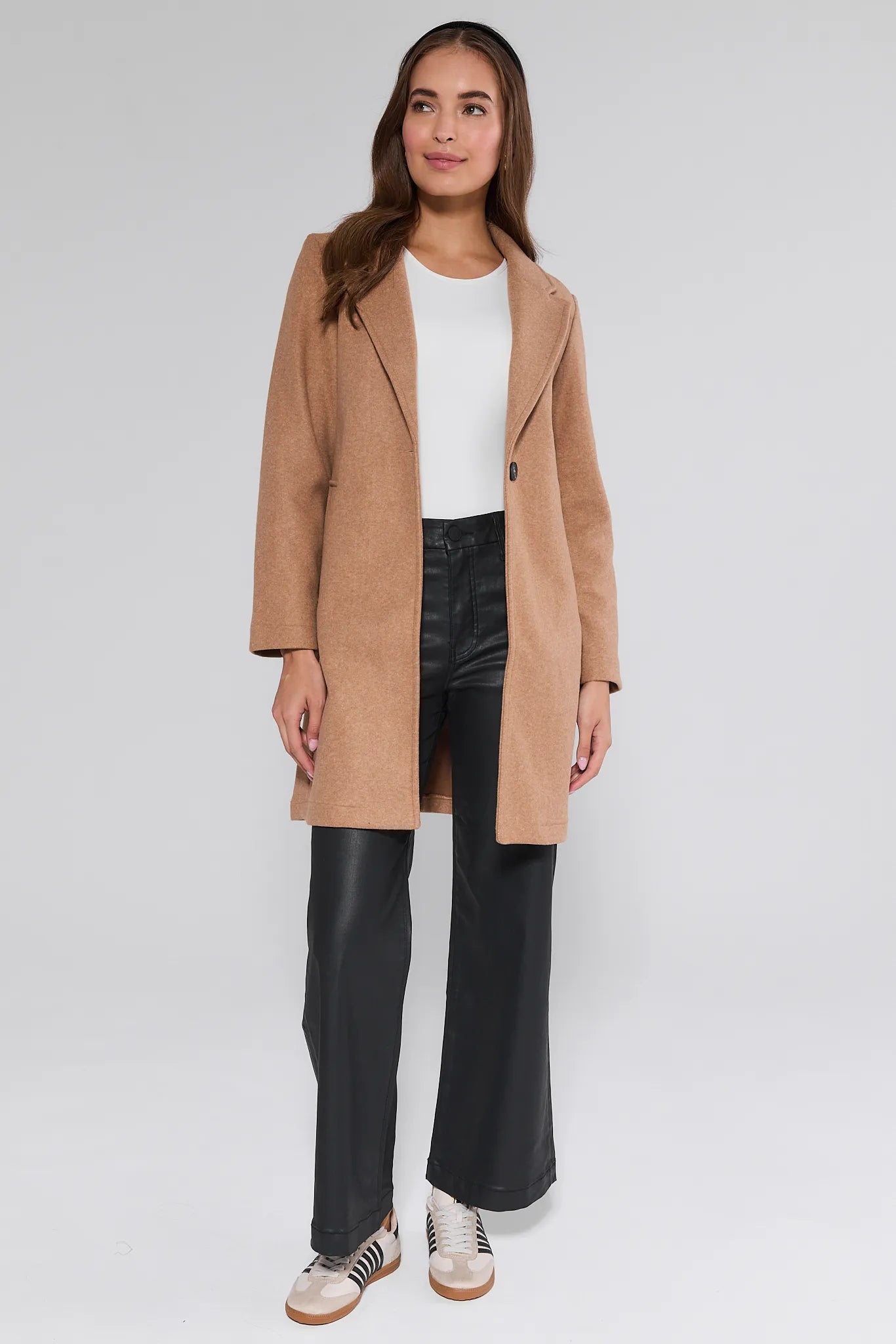 Woman wearing a beige Lauren Coat, white shirt, black pants, and sneakers on a white background
