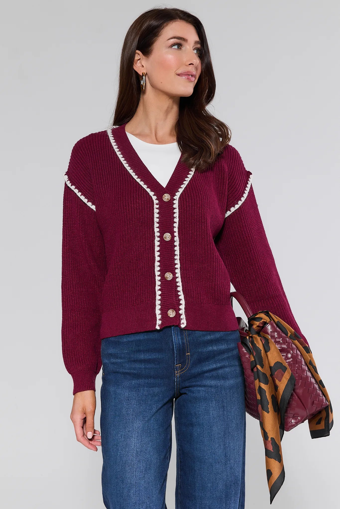 Woman wearing a burgundy Morgan Cardigan with white trim, blue jeans, and holding a patterned bag against a white background.