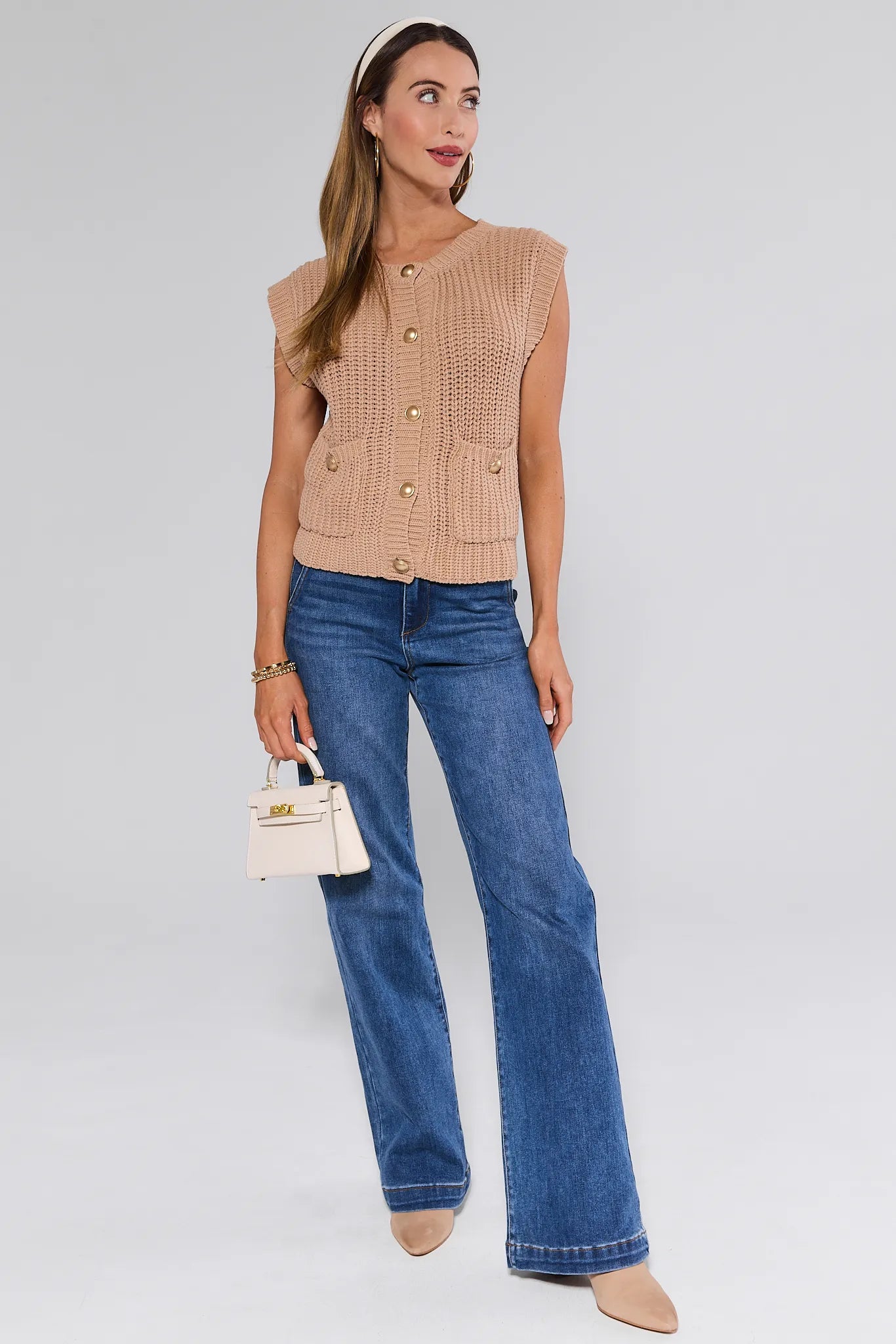Woman wearing a beige knitted Angelina Vest and blue jeans, holding a small handbag on a white background