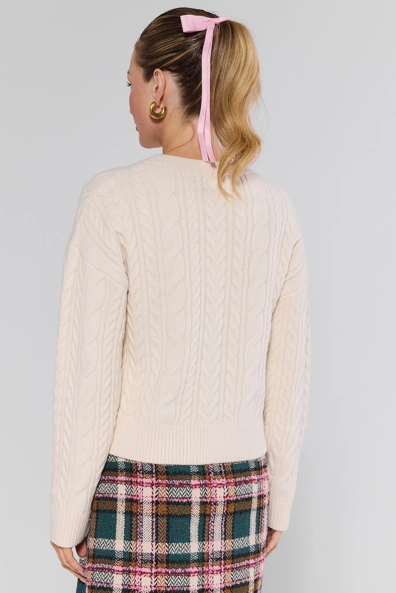 Person wearing a cream cable knit Dakota Sweater and plaid skirt with a pink headband against a gray background