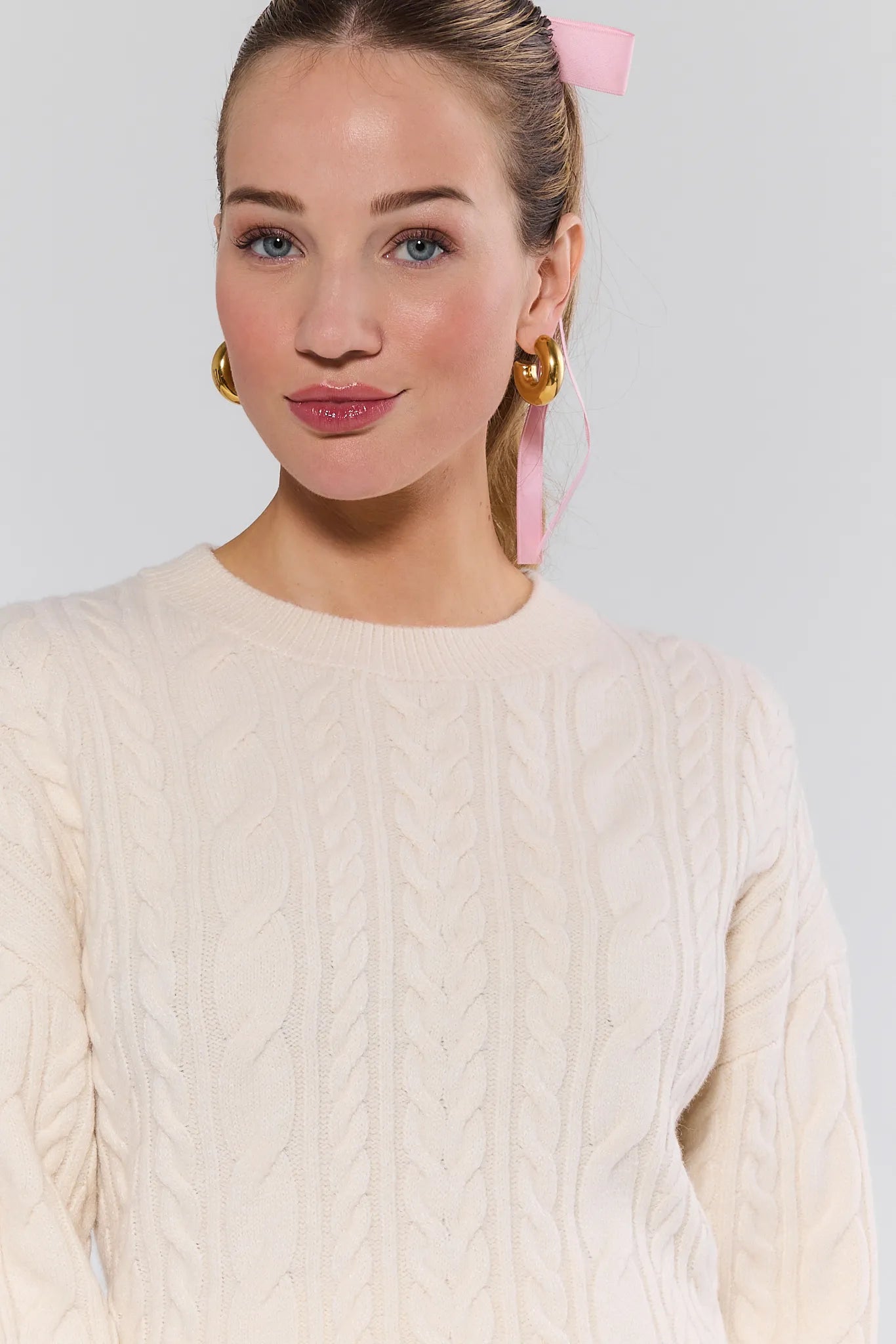 Woman wearing a cream cable knit Dakota Sweater with gold earrings and pink hair accessory on a light gray background
