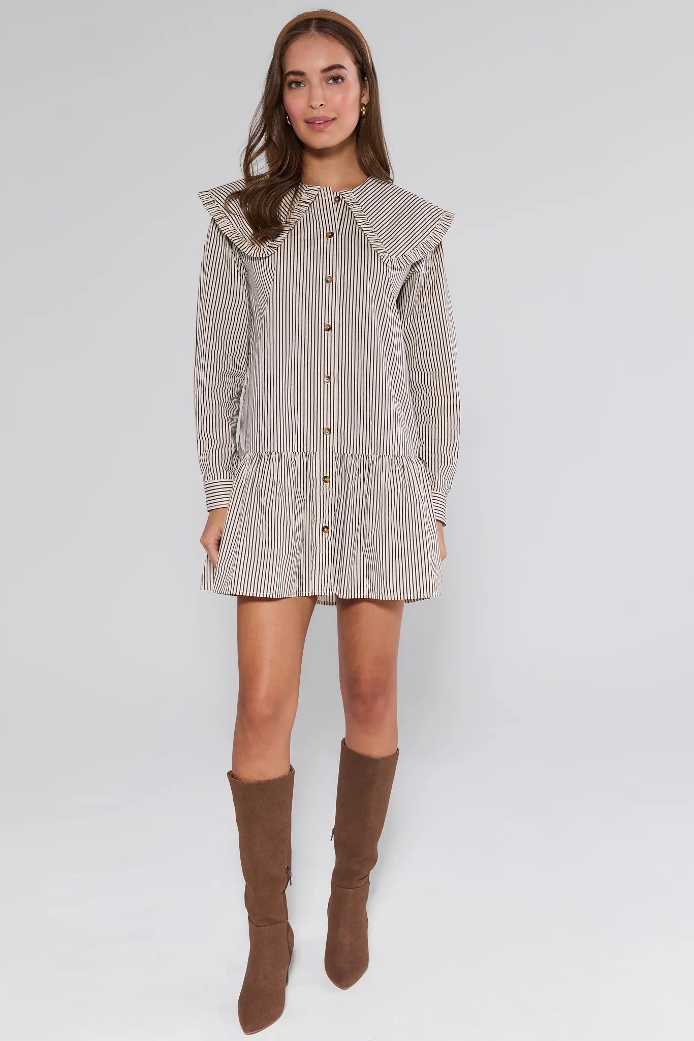 Woman wearing a striped Berkley Dress with ruffled details and brown knee-high boots on a white background