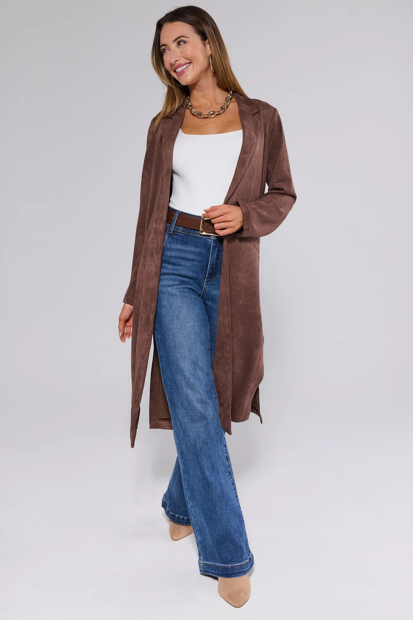 Woman wearing a brown Nadine Coat, white top, and blue jeans on a plain background