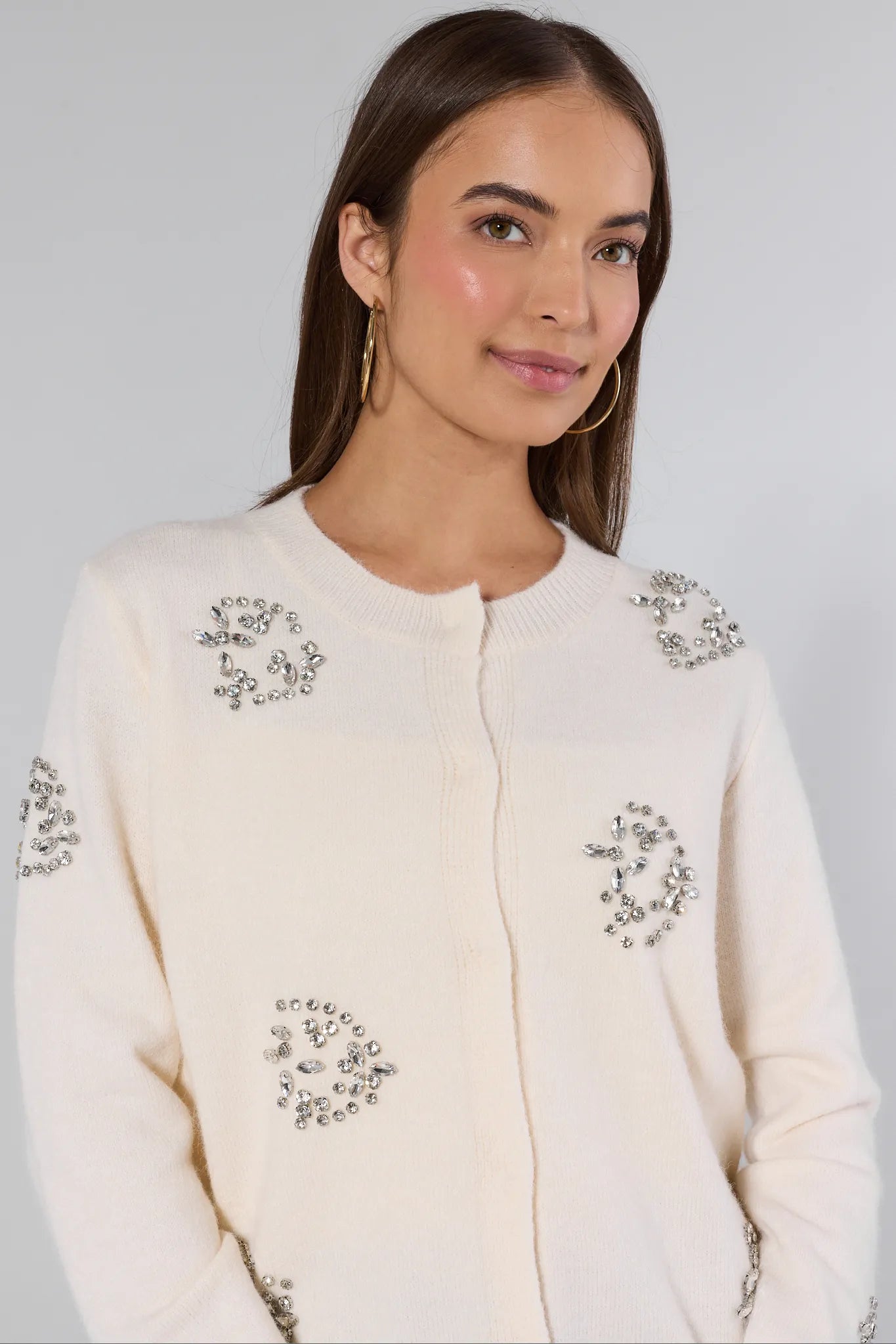 Woman wearing a cream-colored cardigan with decorative embellishments on a plain background