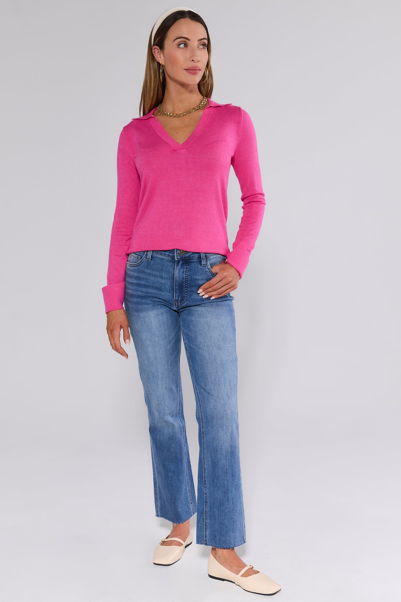 Woman models the Harlen Pink Knit Sweater with a V-neck and collared design, paired with flared jeans, accessorized with gold jewelry and Mary-Jane flats.