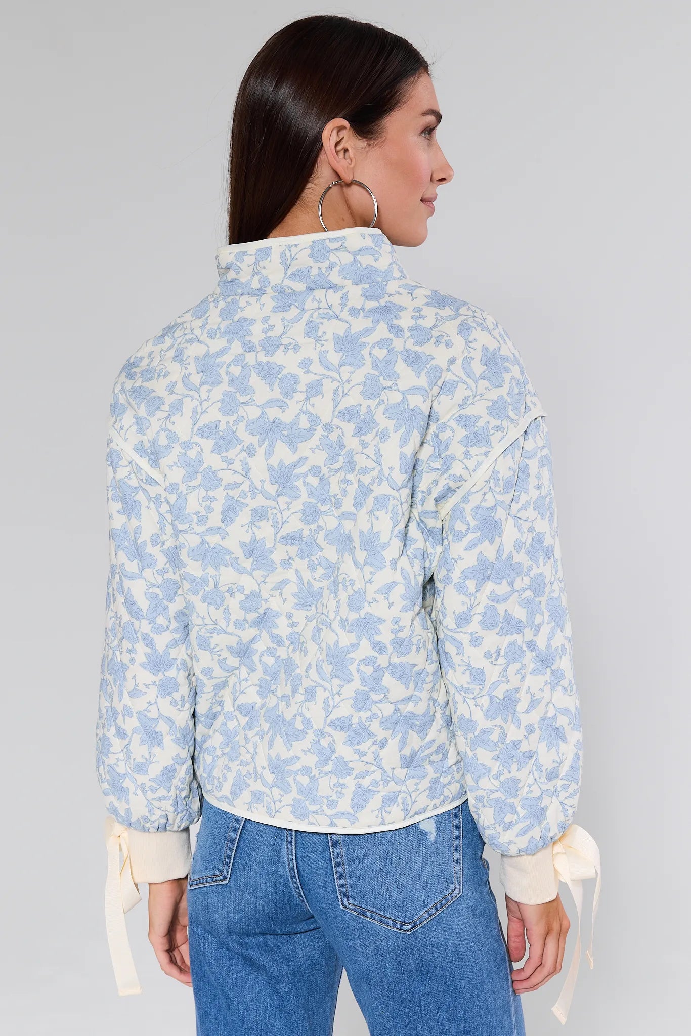 Woman wearing a light blue floral brett jacket over a white shirt with blue jeans on a gray background