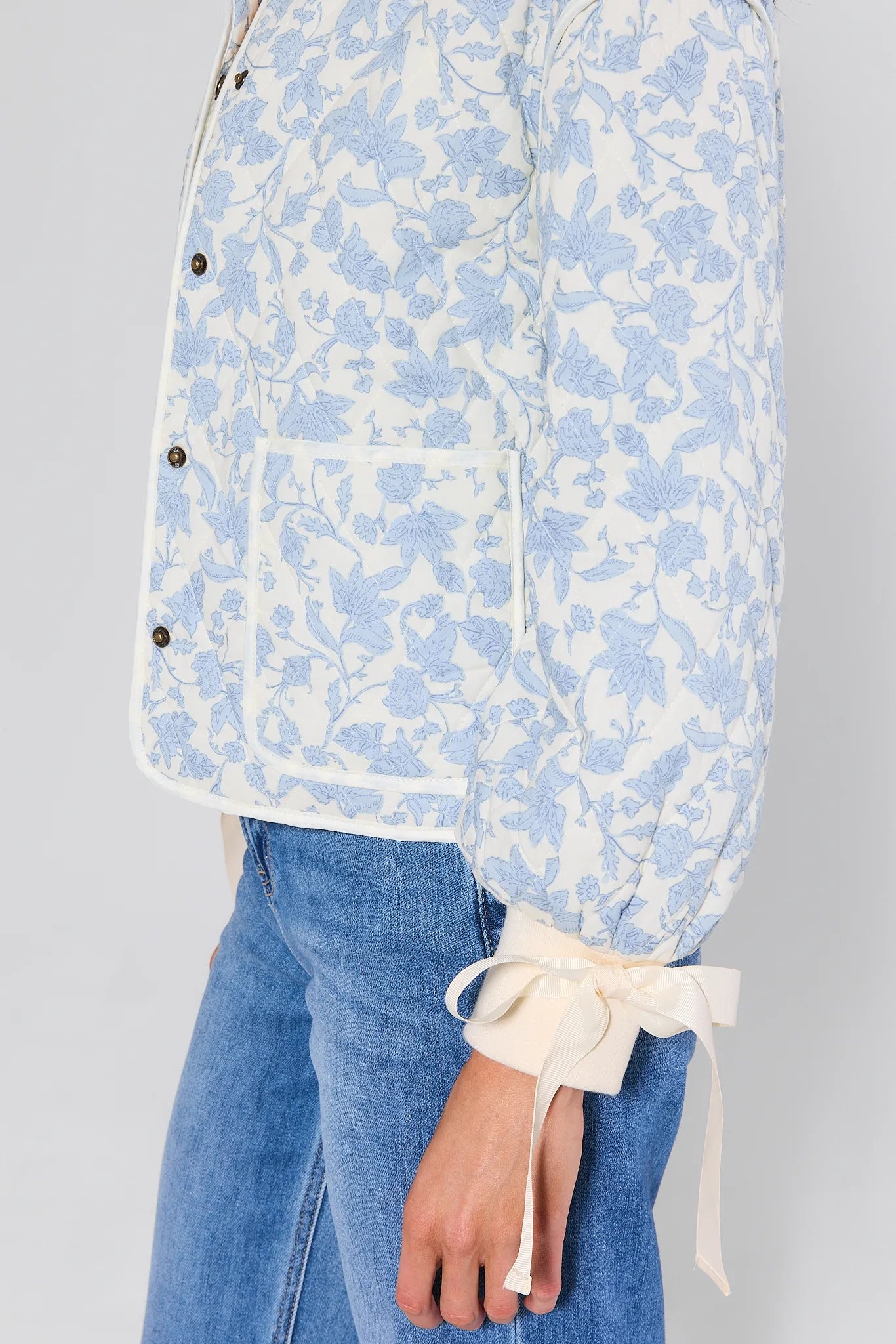 Woman wearing a light blue floral brett jacket over a white shirt with blue jeans on a gray background