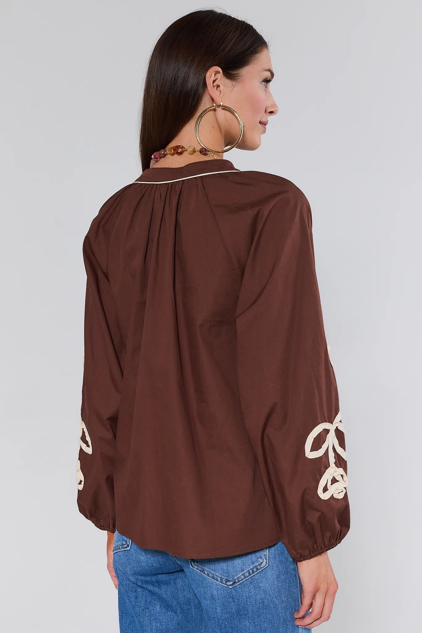 Woman wearing a brown Quinlan Top with white embroidery on sleeves against a white background
