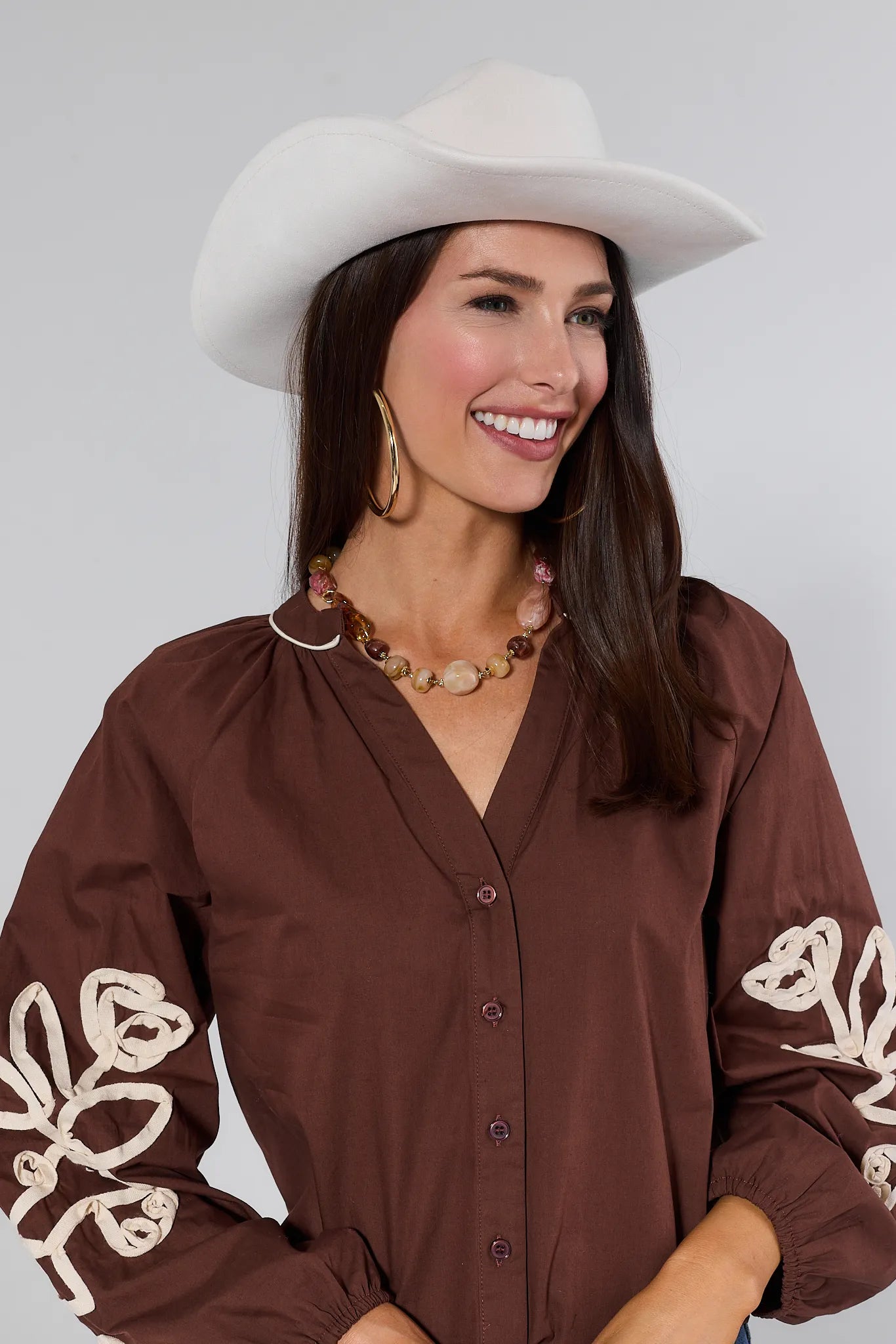 Woman wearing a brown blouse with white embroidery and a white cowboy Lynnie Hat on a plain background