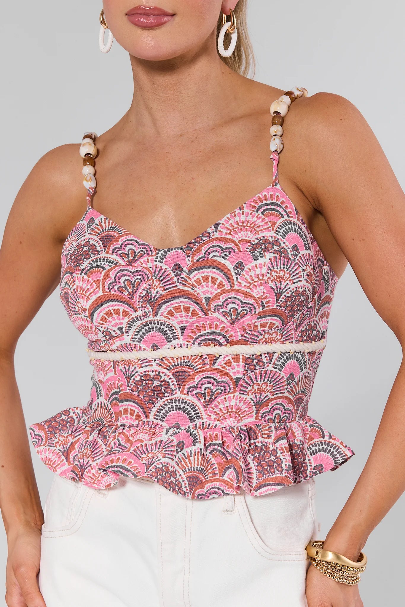 Woman wearing a Susie Pink Printed Peplum Top with beaded straps and white pants on a plain background