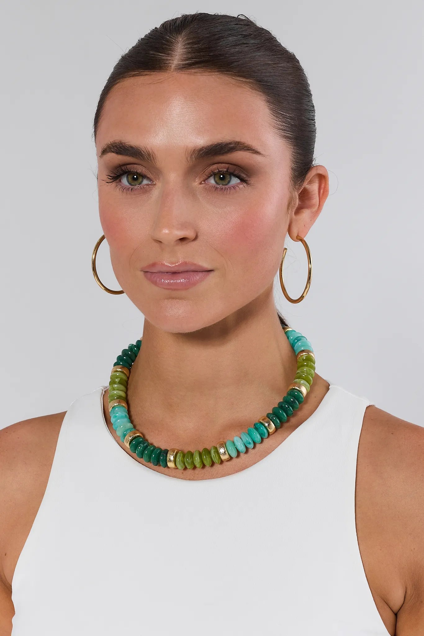 Woman wearing a green beaded necklace and gold hoop earrings on a plain background