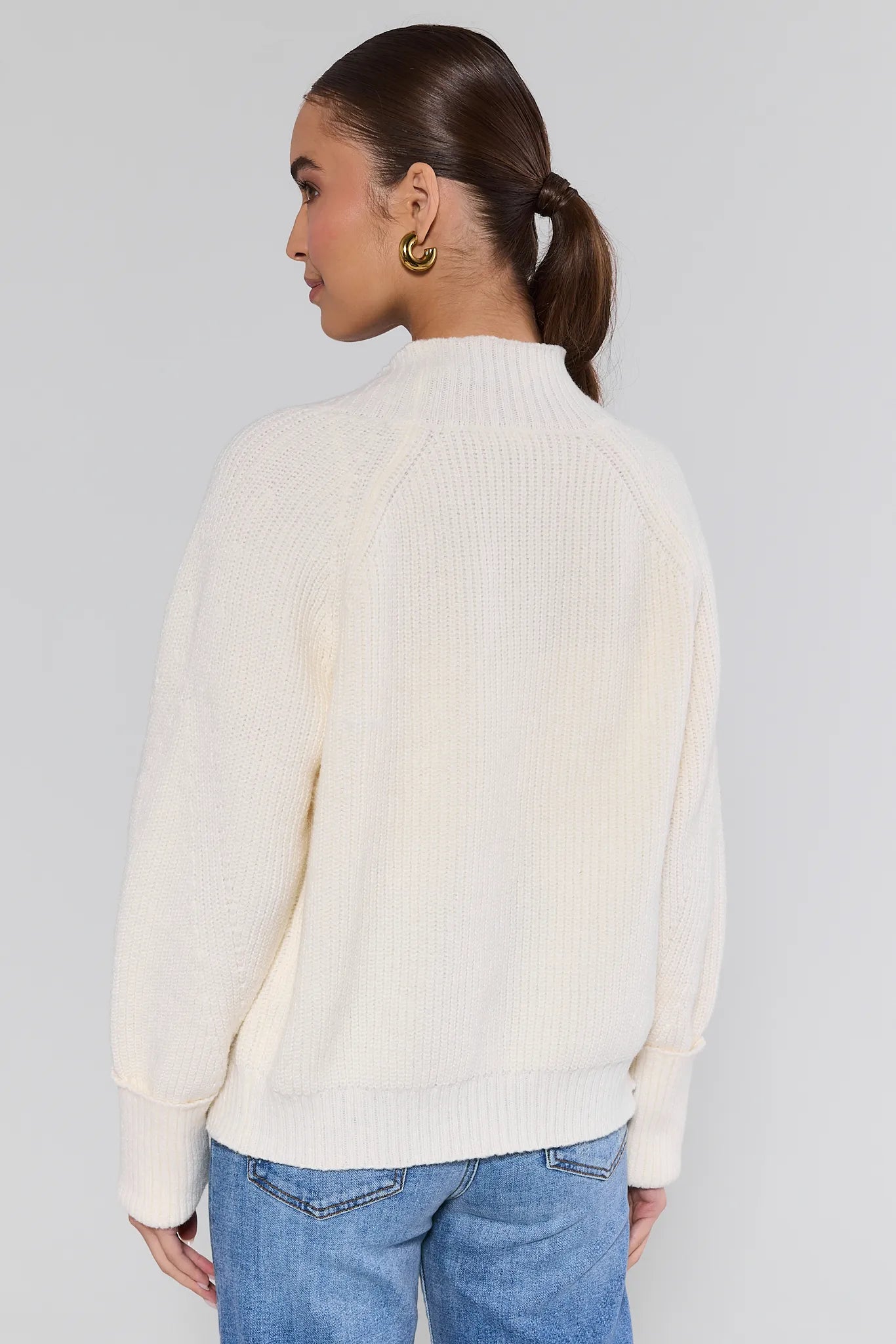 Woman wearing a cream sweater and blue jeans against a plain background