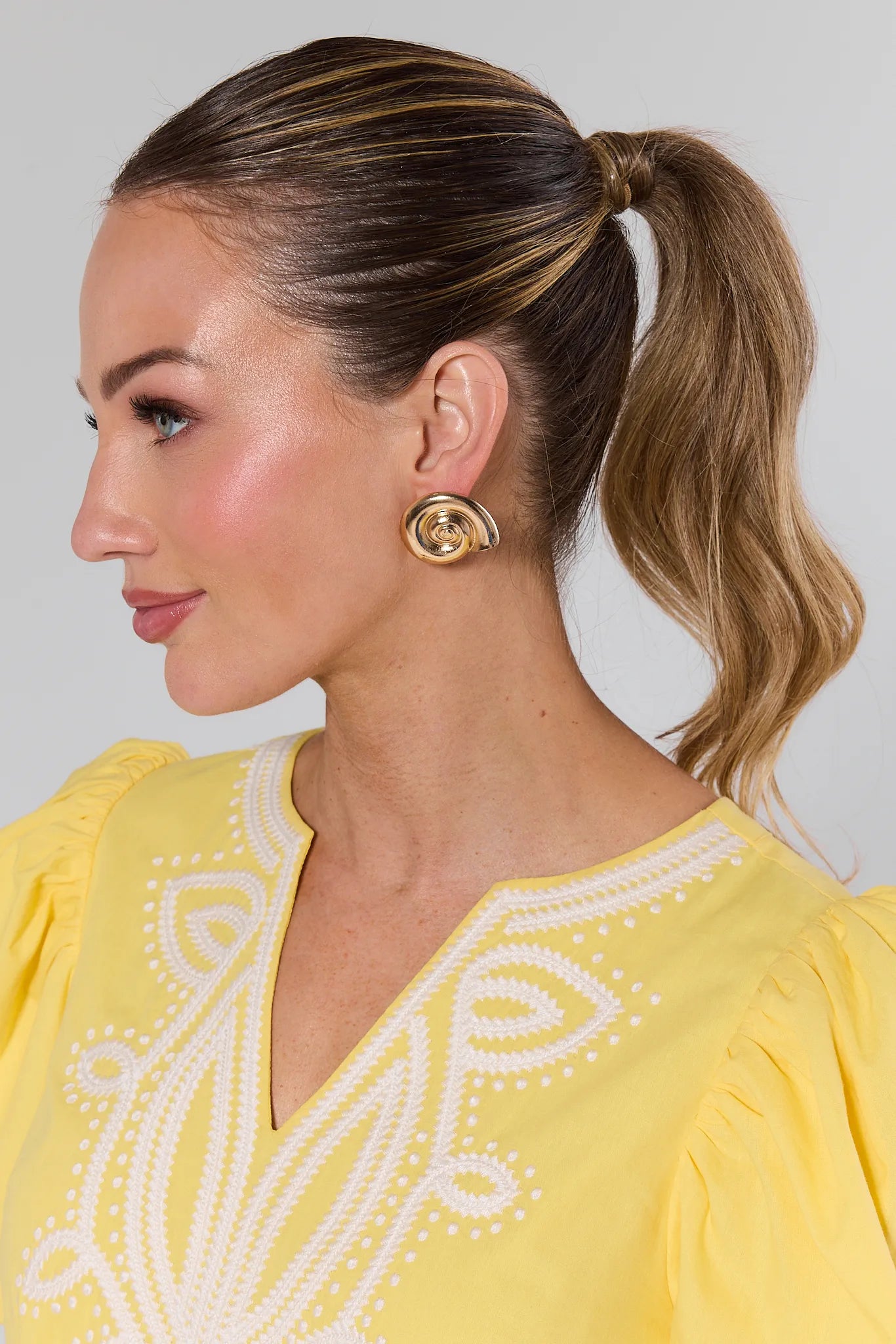 Woman wearing a yellow dress with white embroidery and Mara Gold Seashell Earrings against a plain background