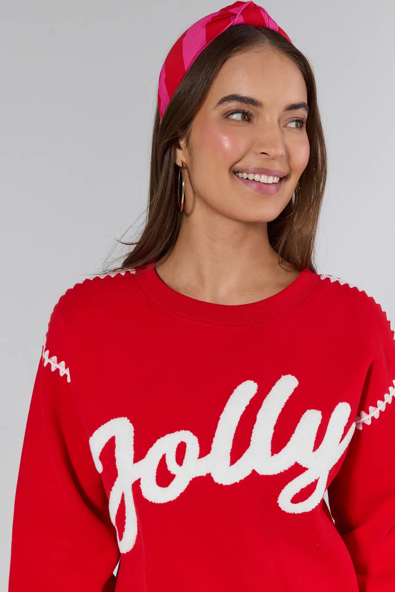 Woman wearing a red sweater with 'Jolly' text and a pink headband against a gray background