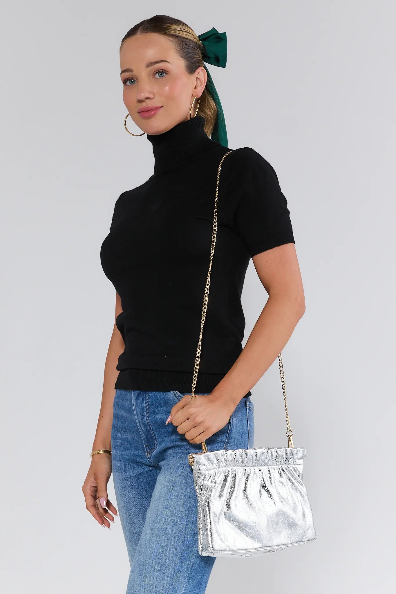 Woman wearing a black turtleneck, blue jeans, and holding a silver handbag against a white background