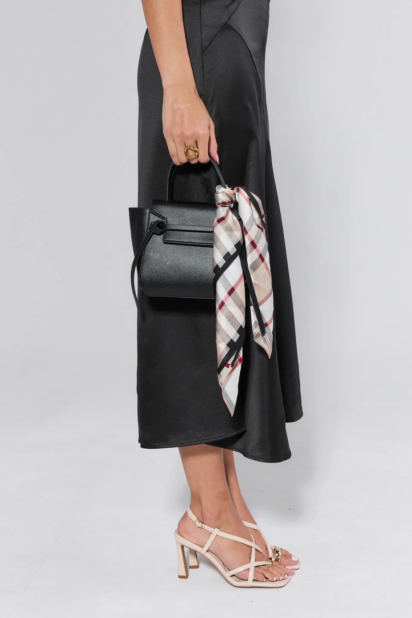 Person holding a black handbag with a plaid scarf, wearing a black dress and beige sandals on a light gray background