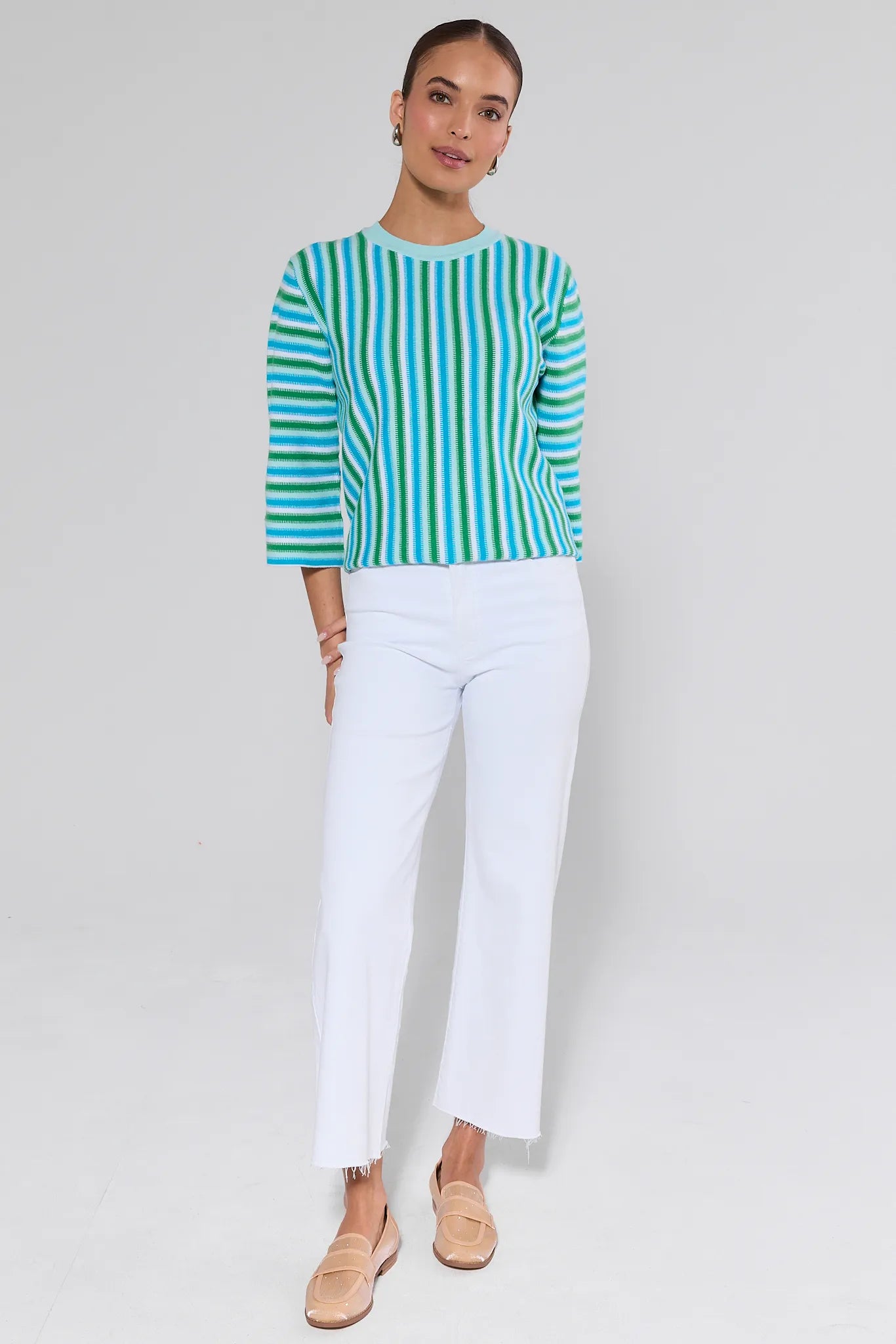Woman wearing a green and blue striped sweater with white pants on a light gray background