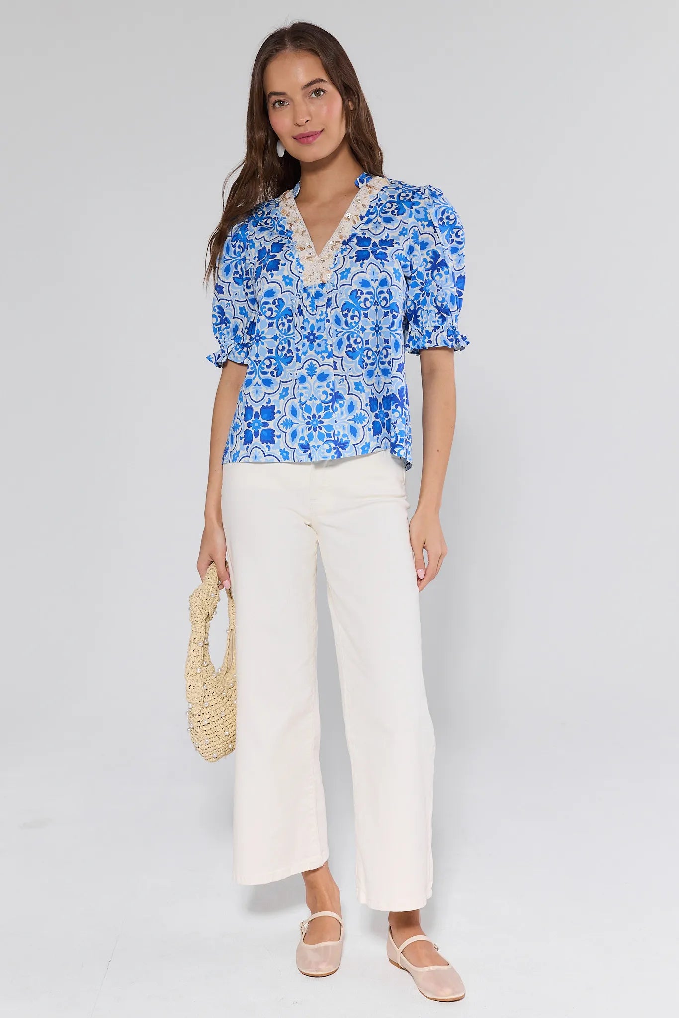 Woman wearing a blue floral blouse and white pants holding a straw bag on a plain background