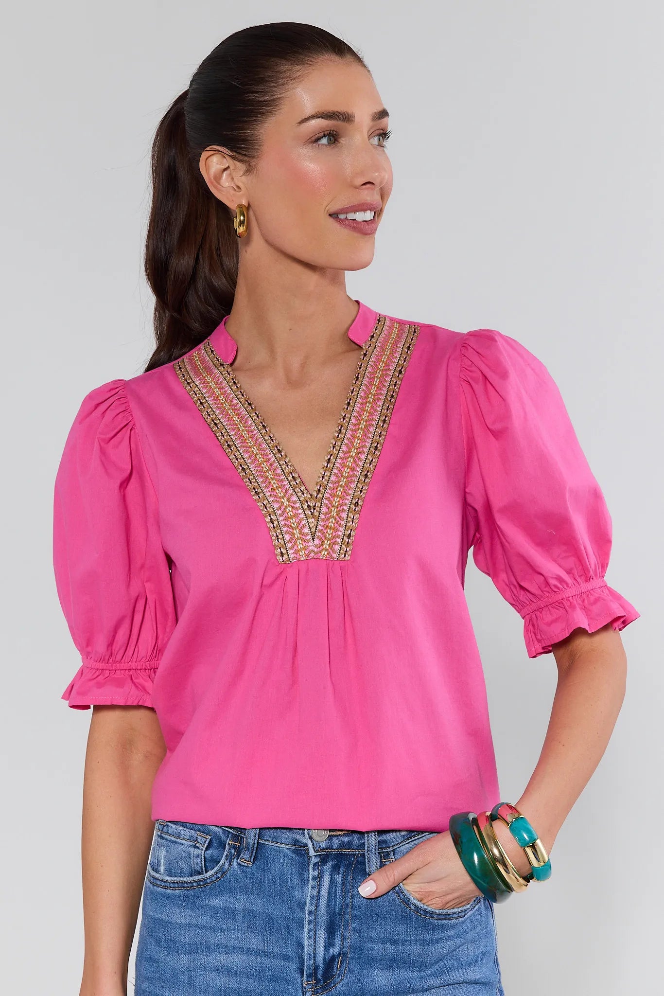 Woman wearing a pink blouse with intricate detailing and blue jeans on a plain background