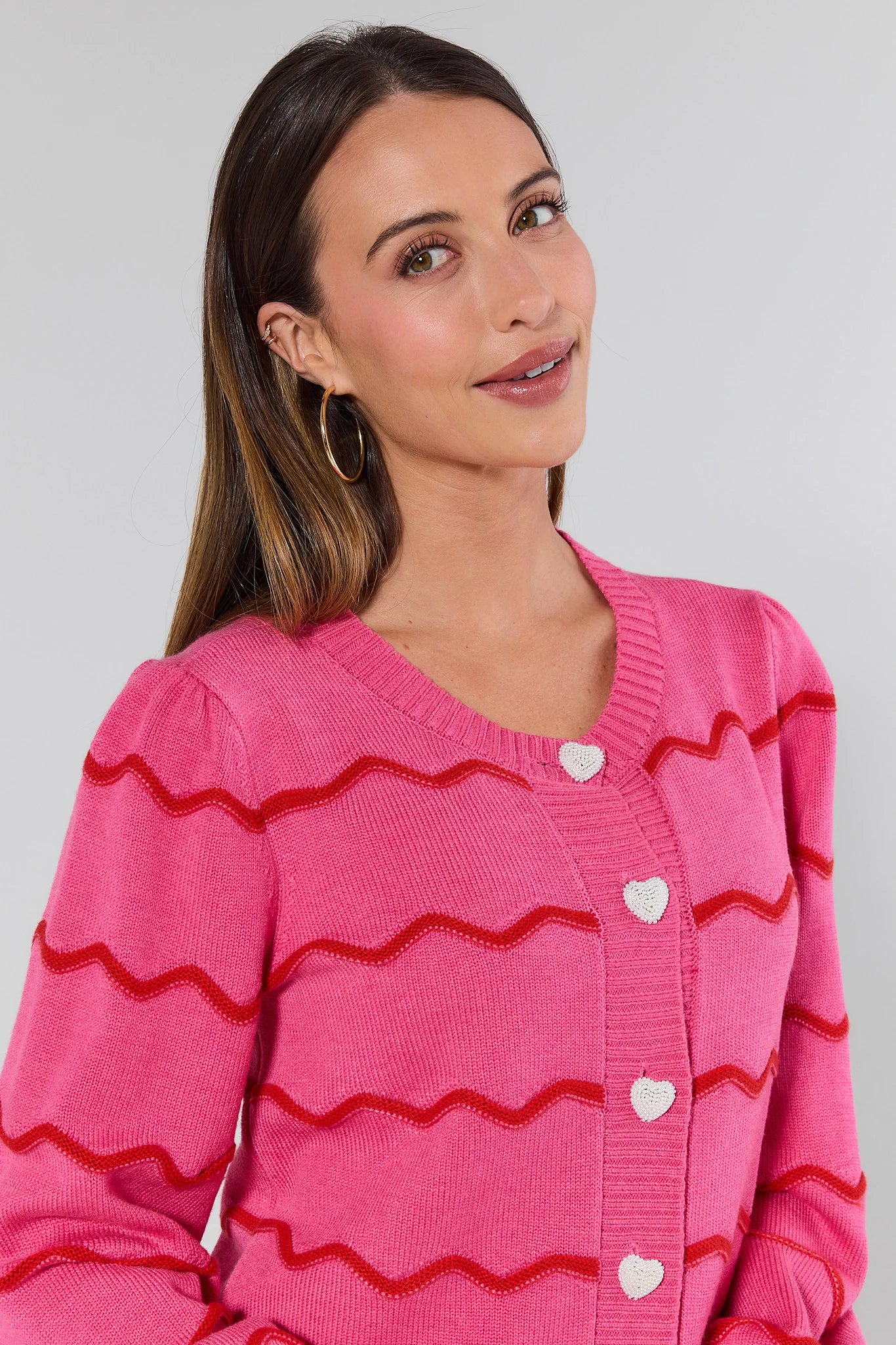 Woman wearing a pink cardigan with red zigzag patterns and white heart buttons on a plain background