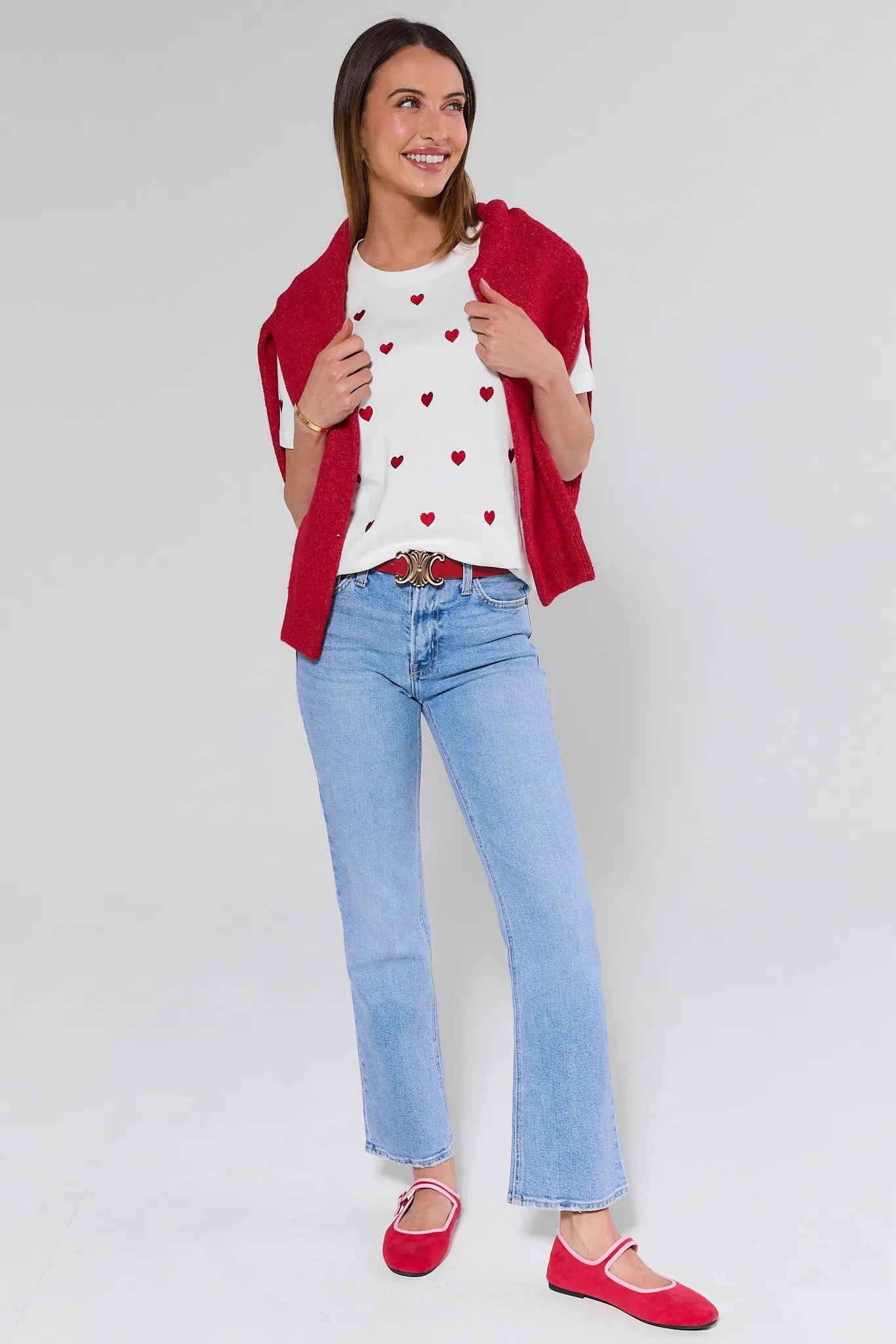 Woman wearing a red cardigan, white shirt with heart patterns, blue jeans, and red shoes on a white background