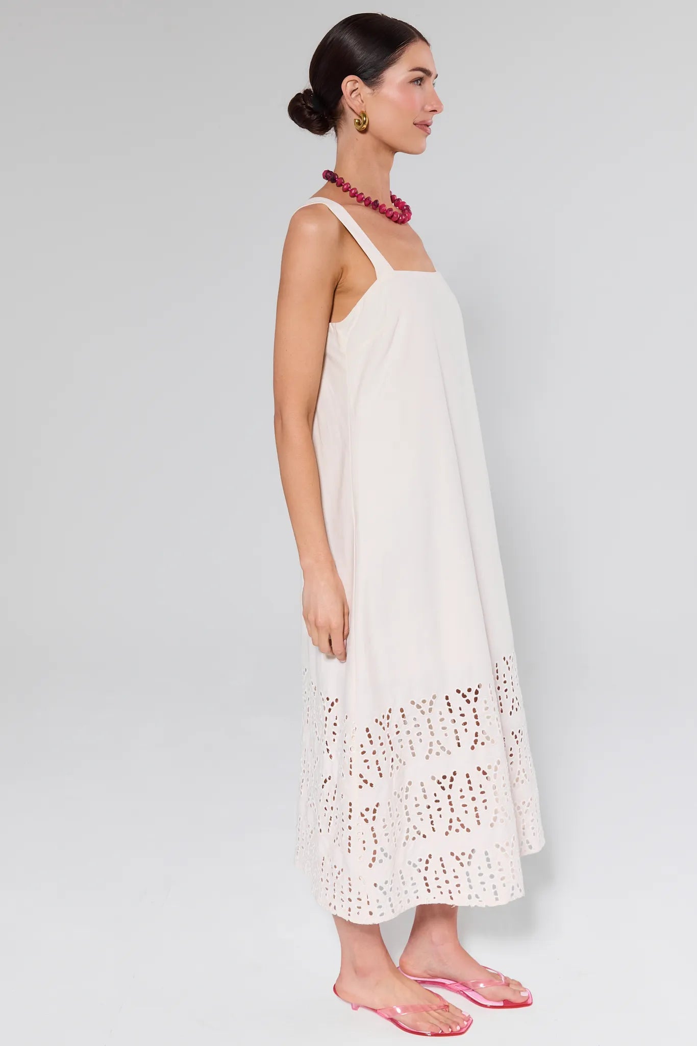 Woman modeling the Melody Bone Eyelet Midi Dress, featuring a tiered skirt, sleeveless design, square neckline, and eyelet embroidery, accessorized with fuchsia necklace and pink sandals.