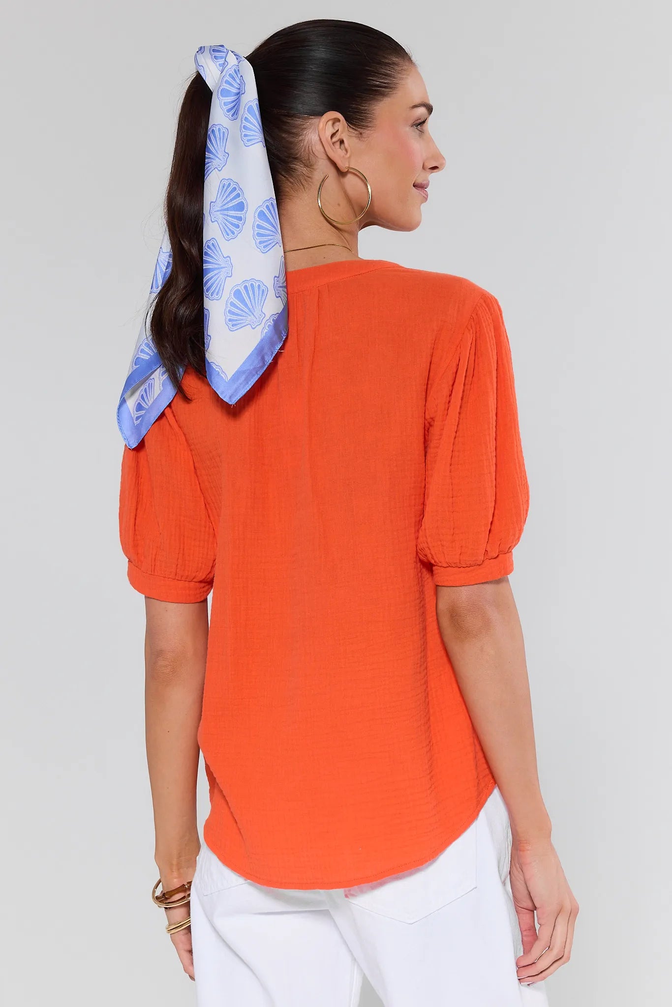 Woman wearing an orange top with puff sleeves and a blue headscarf with white patterns on a plain background