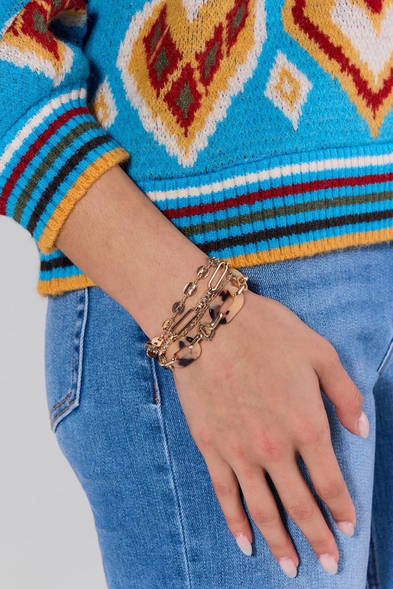 Close-up of a hand wearing gold bracelets on a person wearing a colorful sweater and blue jeans.